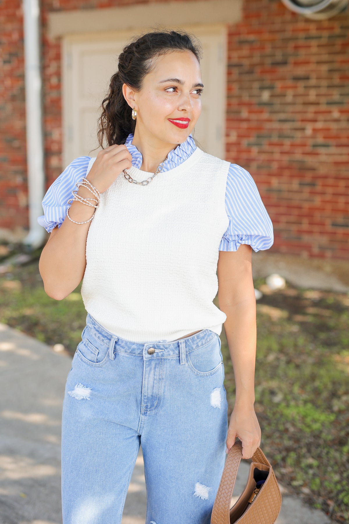 A woman wearing a white knit sweater vest over a light blue and white striped short-sleeve blouse, paired with light-wash distressed jeans, standing outdoors in front of a brick building.
