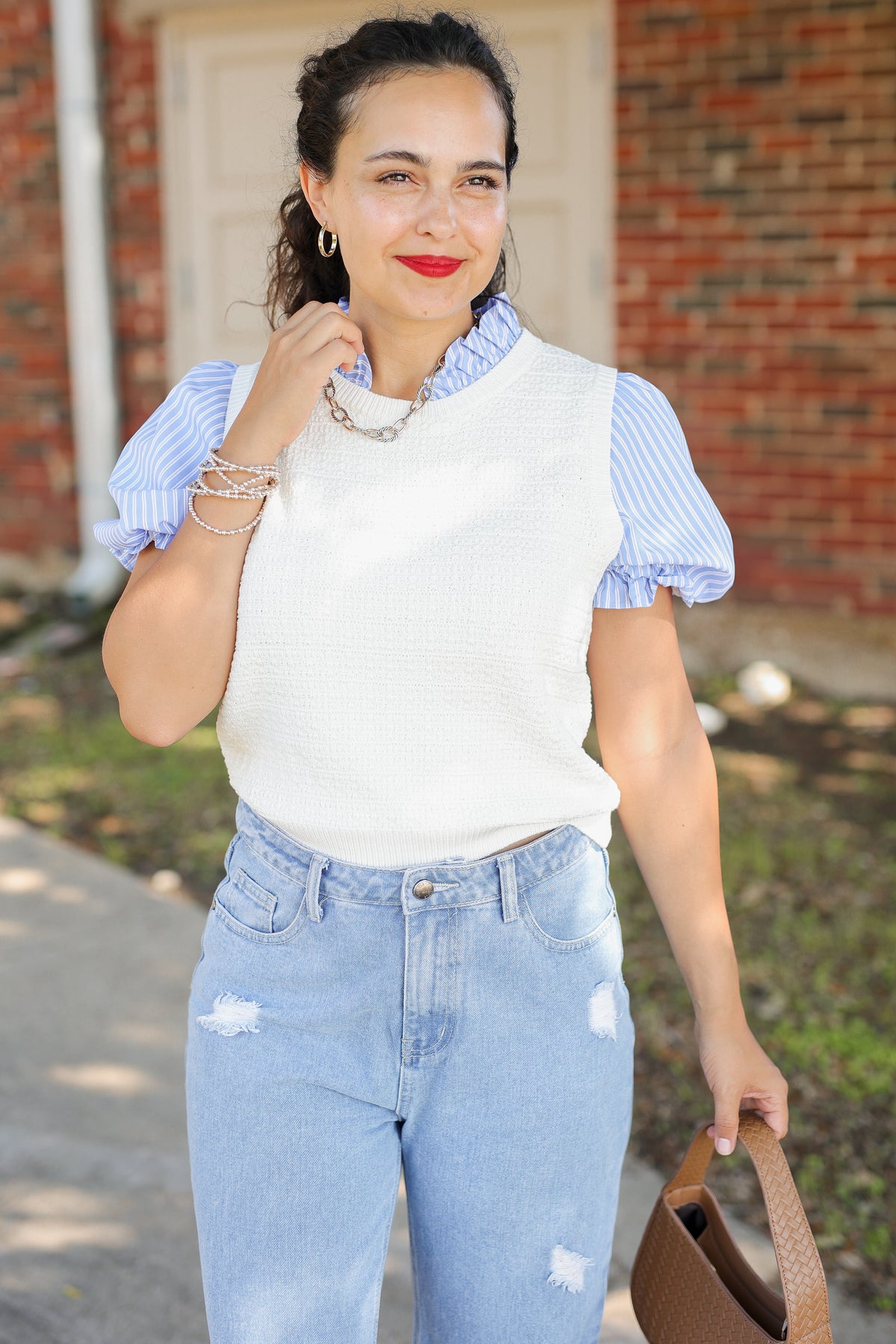A woman wearing a white ribbed knit vest over a light blue and white striped short-sleeve blouse, paired with light-wash distressed jeans, standing outdoors in front of a brick building.