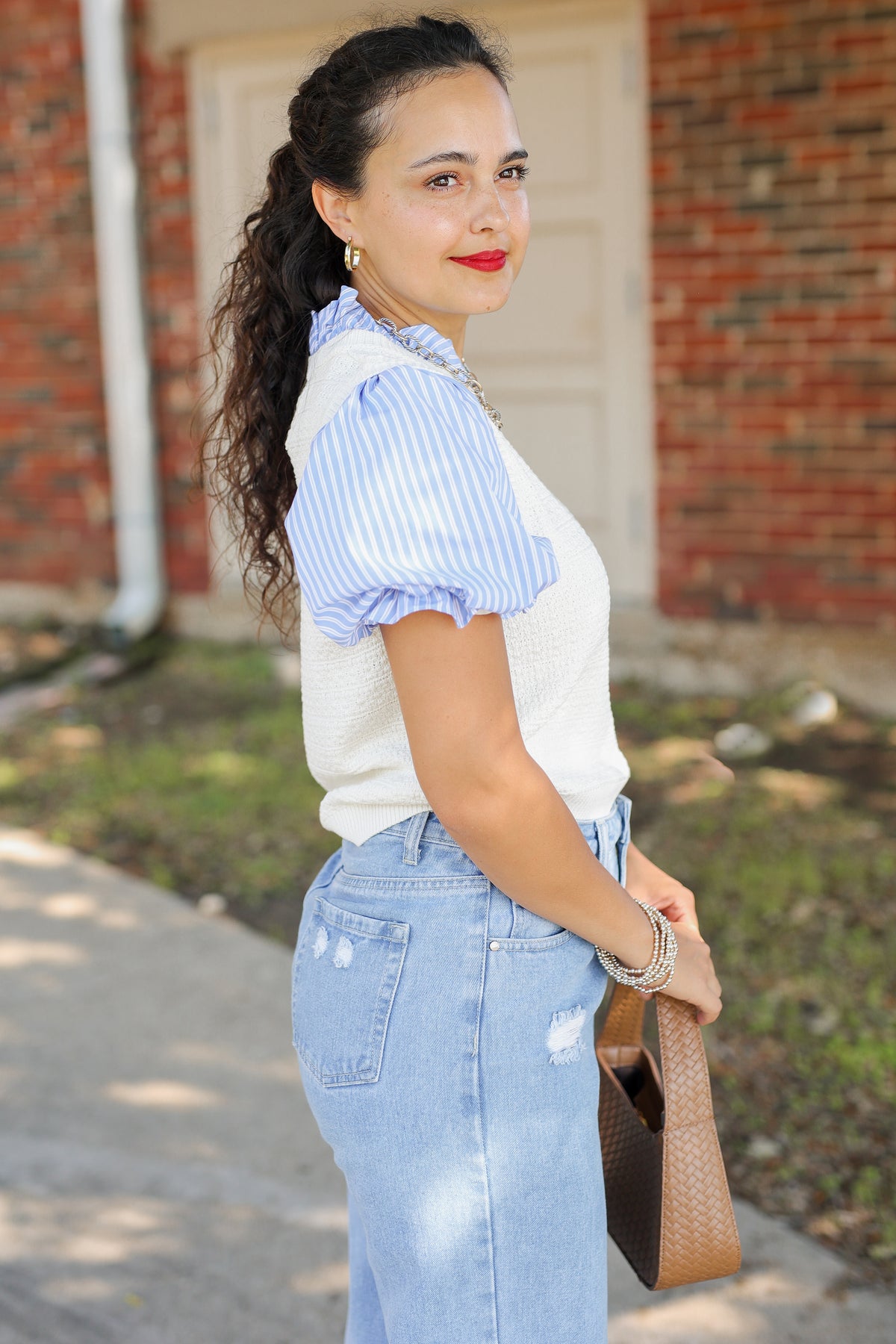 A woman wearing a white ribbed knit top with light blue and white vertically striped puff sleeves, paired with light blue high-waisted jeans, holding a brown woven handbag outdoors in front of a brick building.