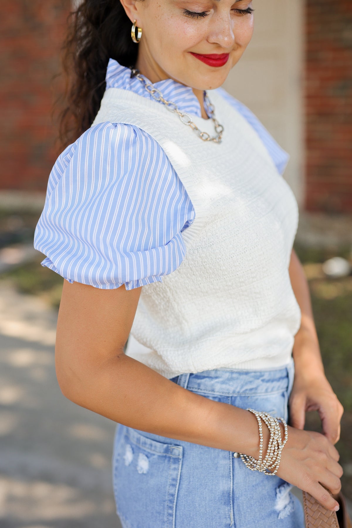 A woman wearing a white textured knit vest over a light blue and white striped puff-sleeve blouse, paired with light-wash distressed denim jeans and layered gold bracelets.