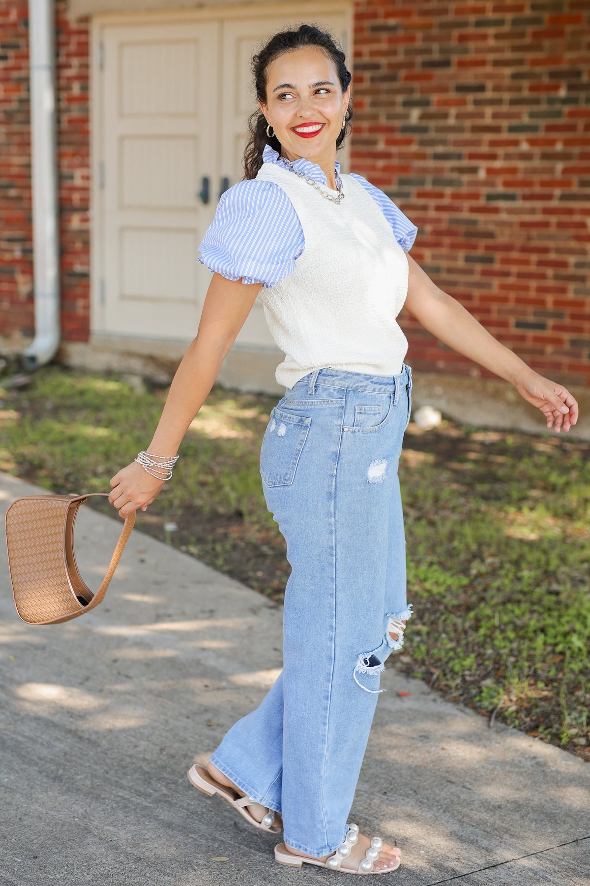 A woman stands outdoors on a sidewalk in front of a brick building, wearing a white sleeveless vest over a light blue and white striped short-sleeve blouse, light-wash distressed jeans, and flat sandals. She is holding a woven tan handbag.