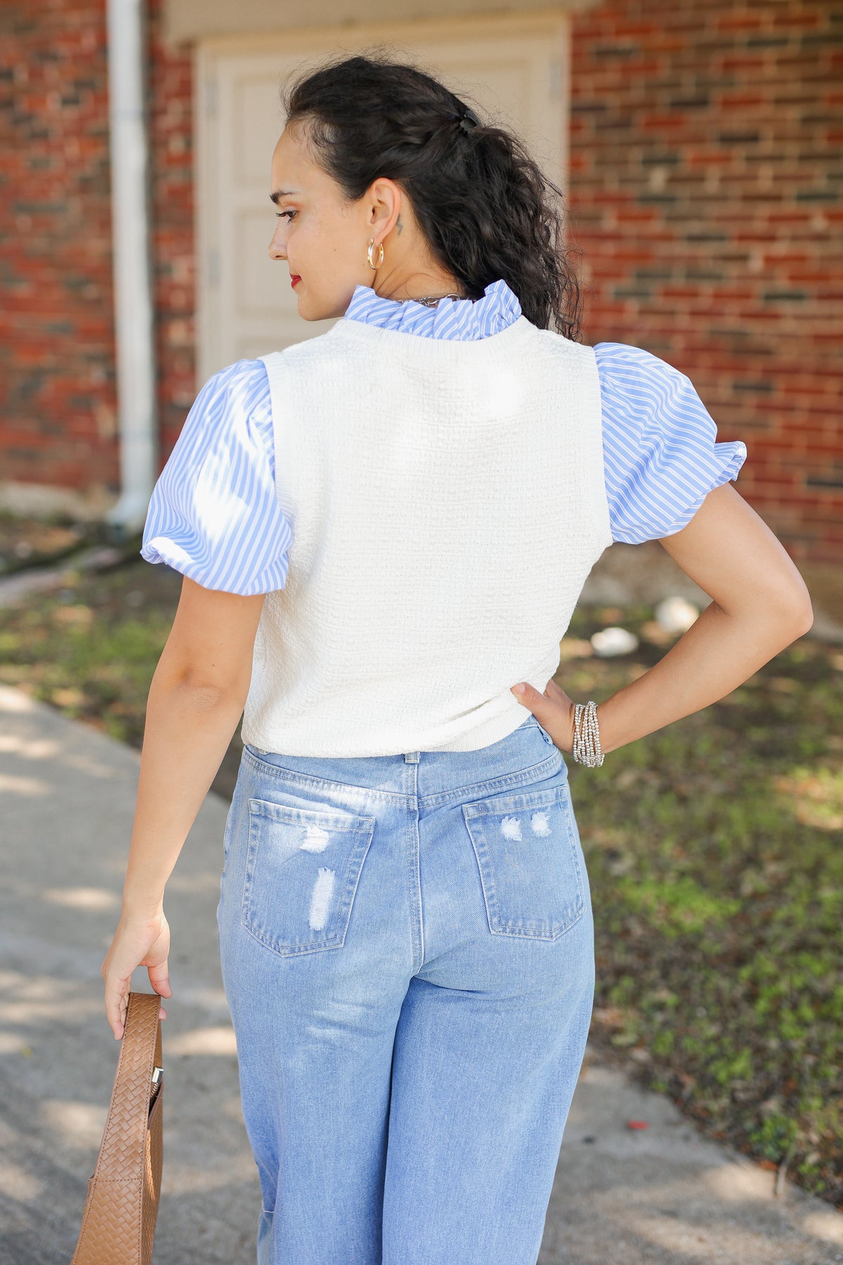 A woman is shown from behind wearing a white knit vest over a light blue and white striped puff-sleeve blouse, paired with light-wash blue jeans, standing outdoors in front of a brick building.