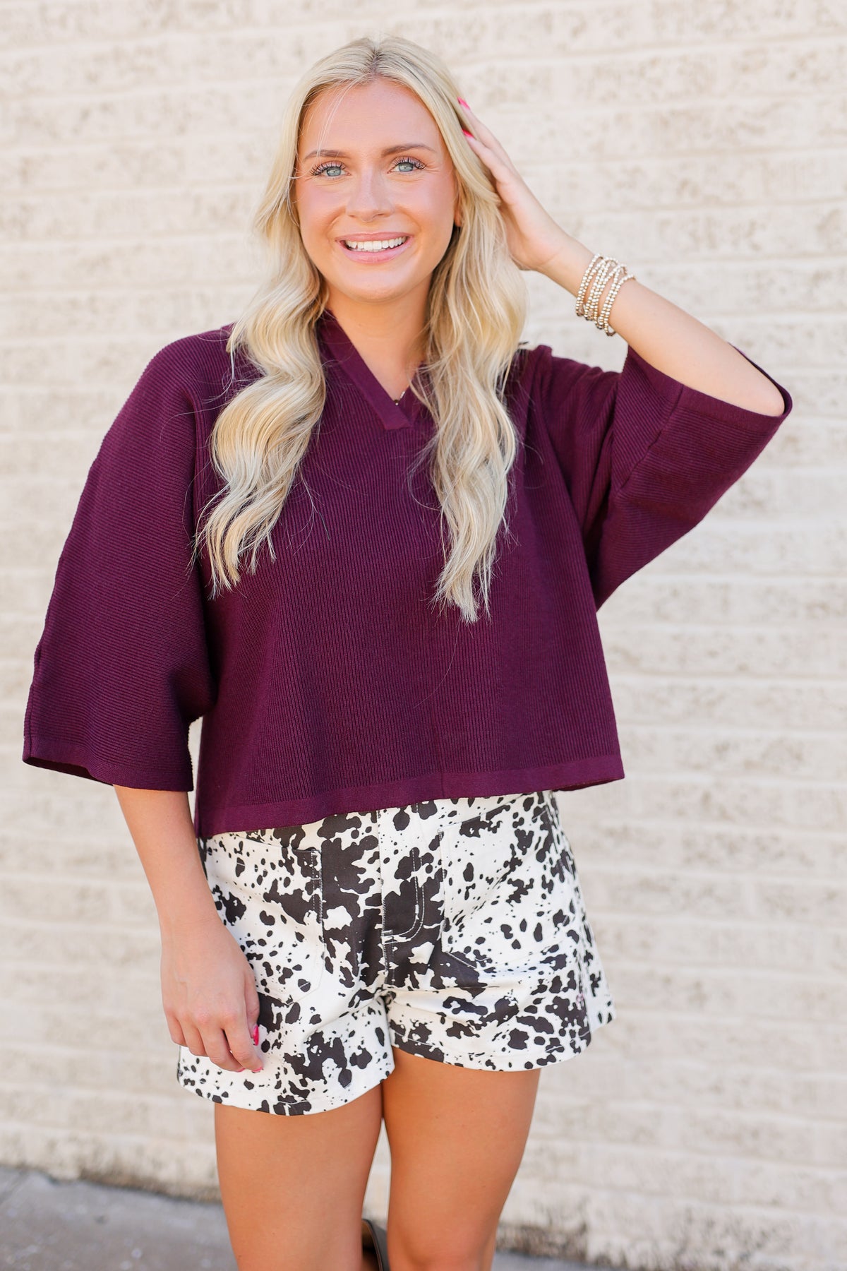 A smiling blonde woman wearing a loose, oversized deep plum-purple short-sleeve top and black-and-white patterned shorts, standing against a light beige brick wall.