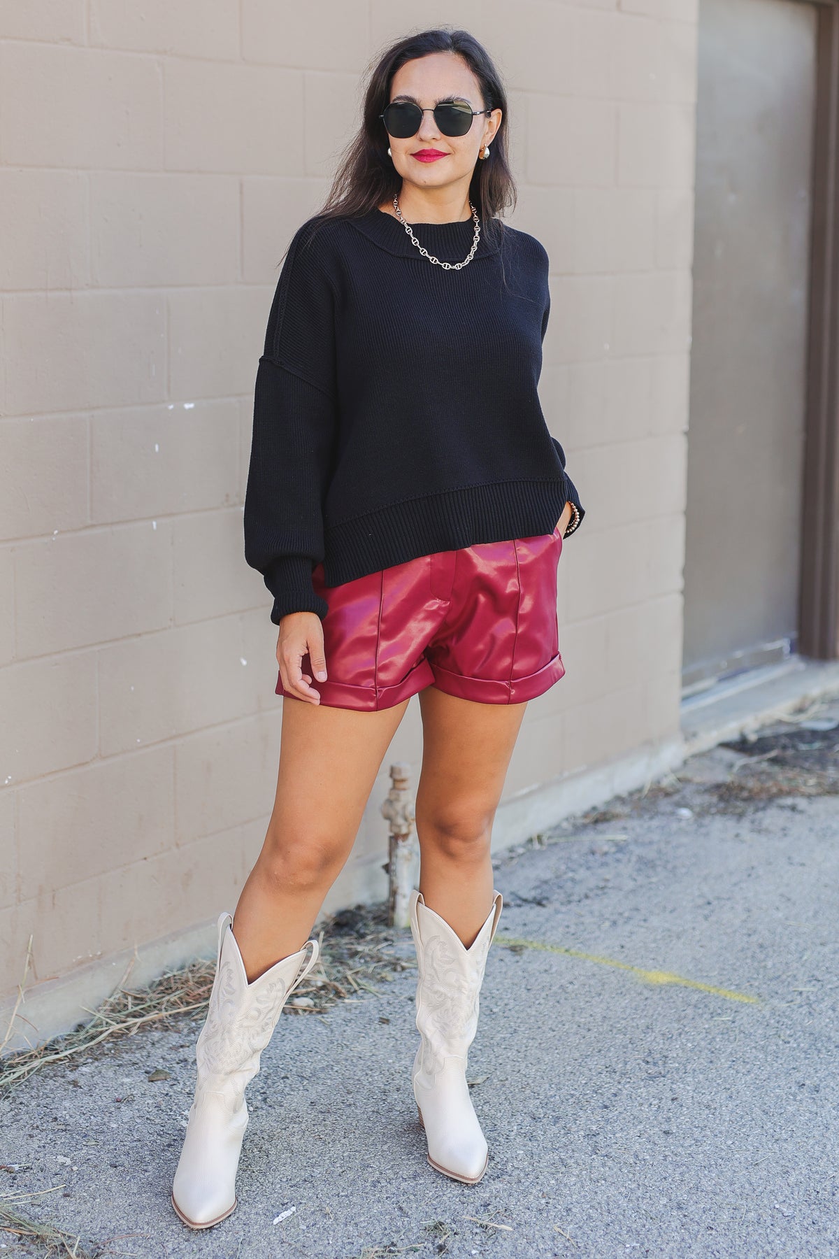 Woman wearing a black sweater, red shorts, and white boots against a beige wall.