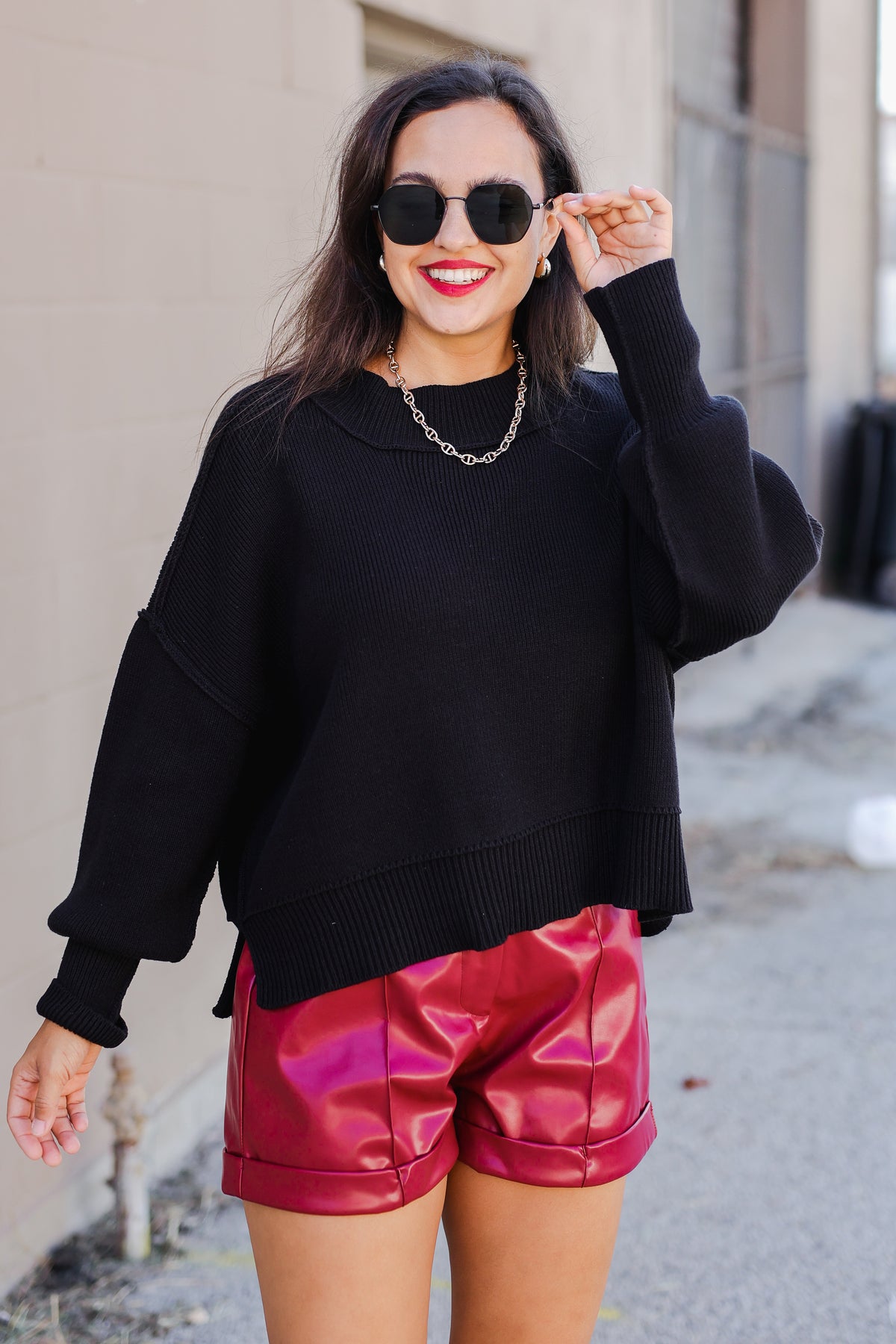 Woman wearing a black sweater and red shorts with sunglasses, standing against a neutral background.