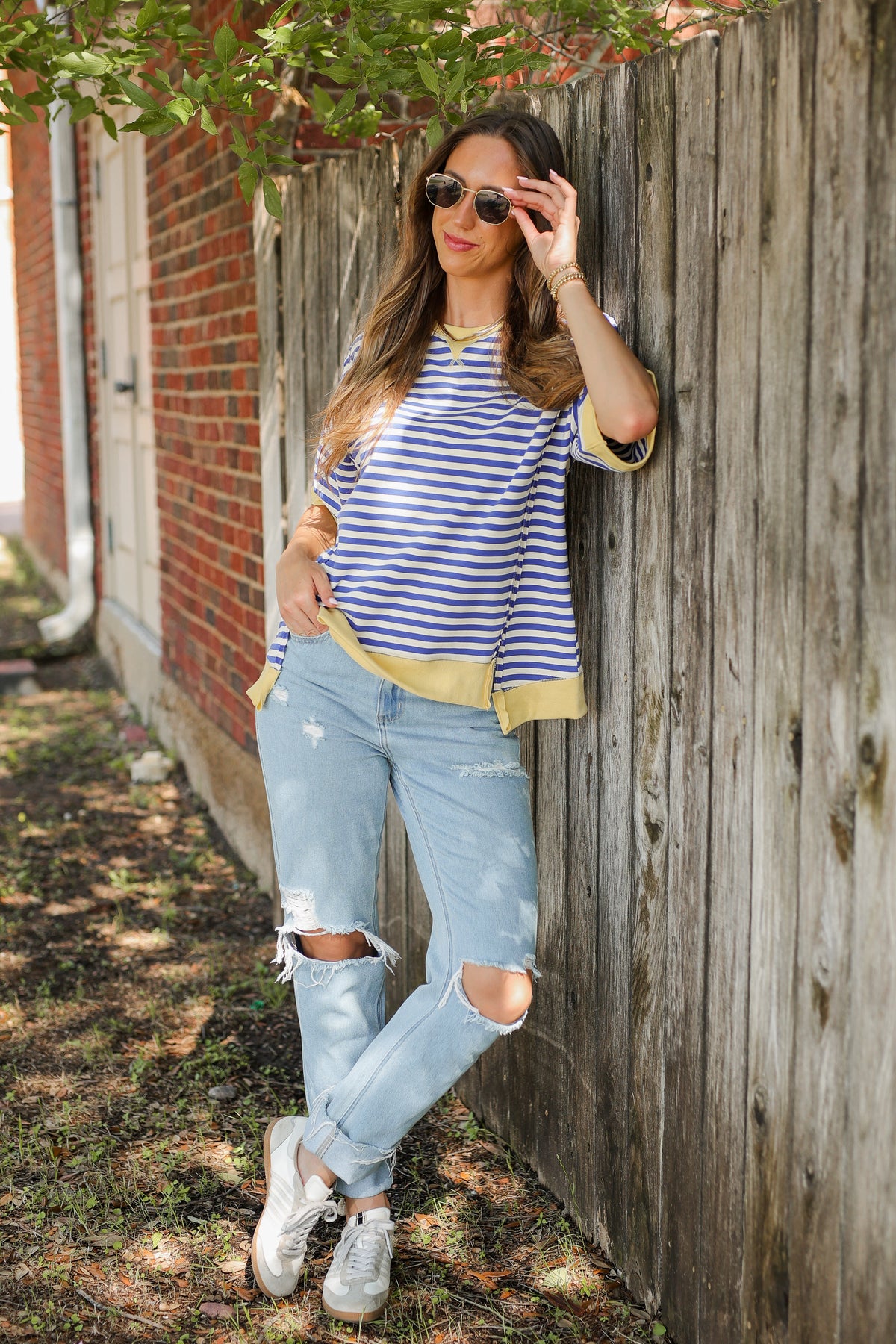 Woman in striped shirt and ripped jeans leaning against a wooden fence.