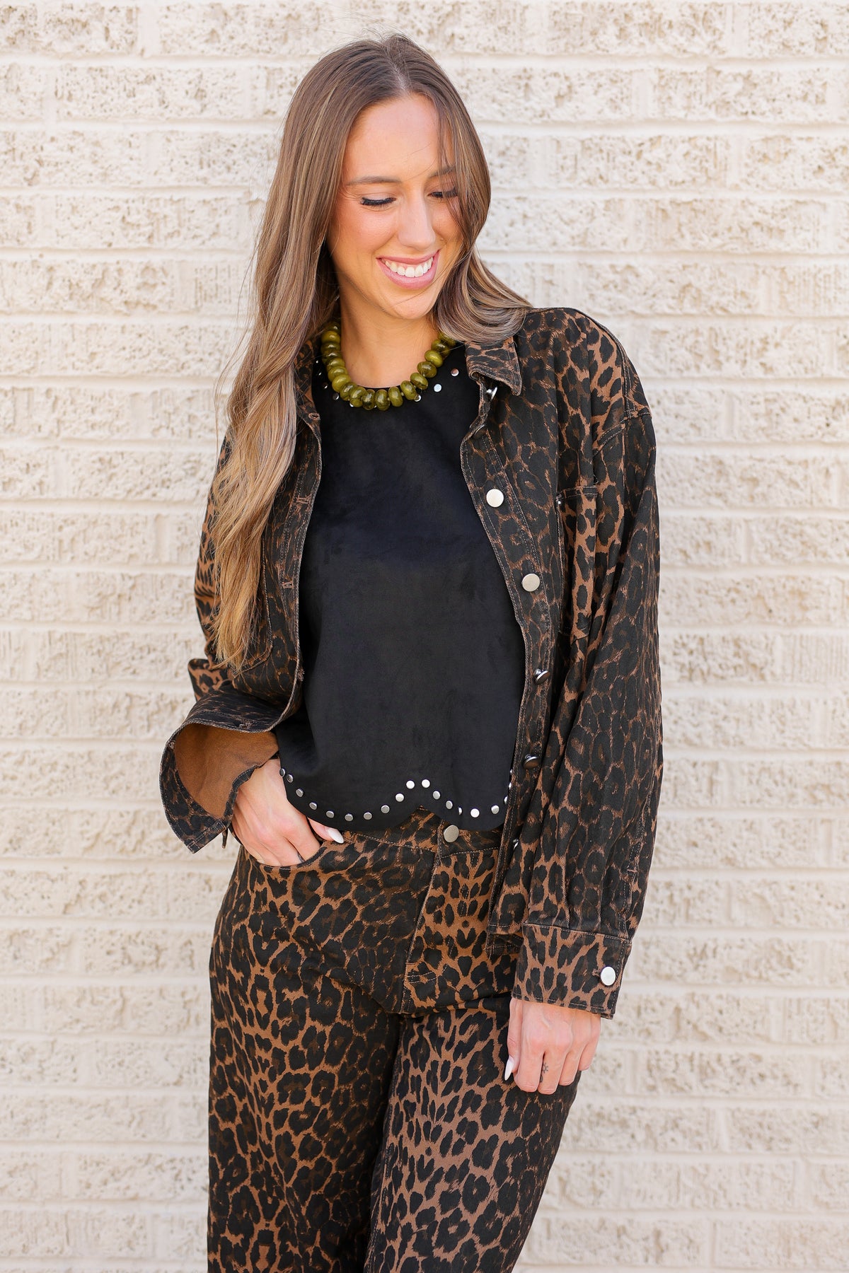 A woman modeling a matching leopard print jacket and pants set with a black top, standing against a light beige brick wall.