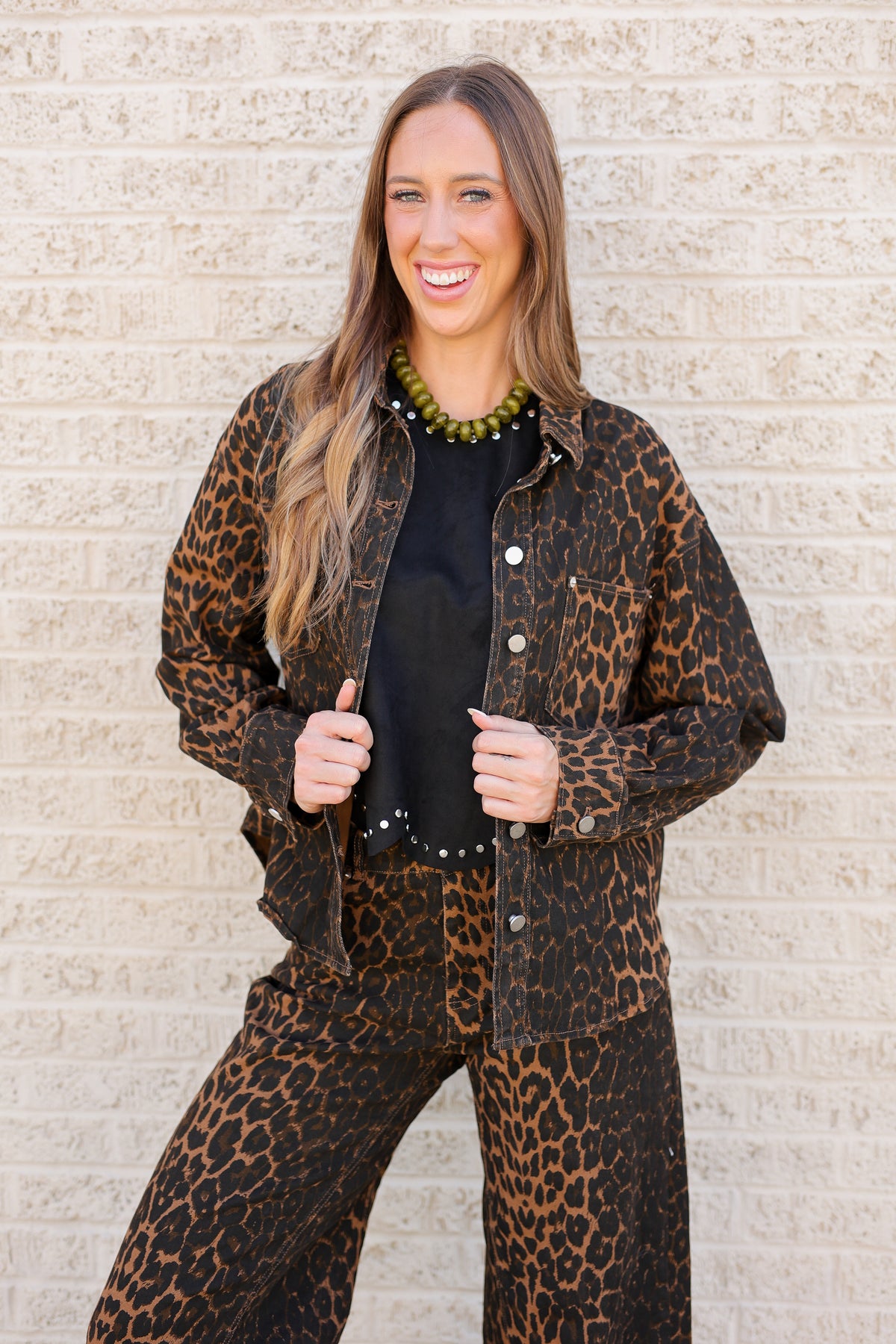 A woman modeling a matching leopard-print jacket and wide-leg pants set with a black top and green beaded necklace, standing against a light beige brick wall.