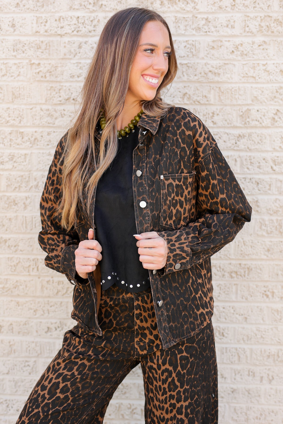 A woman modeling a matching leopard-print button-up shirt and pants set over a black top, standing against a light beige brick wall.
