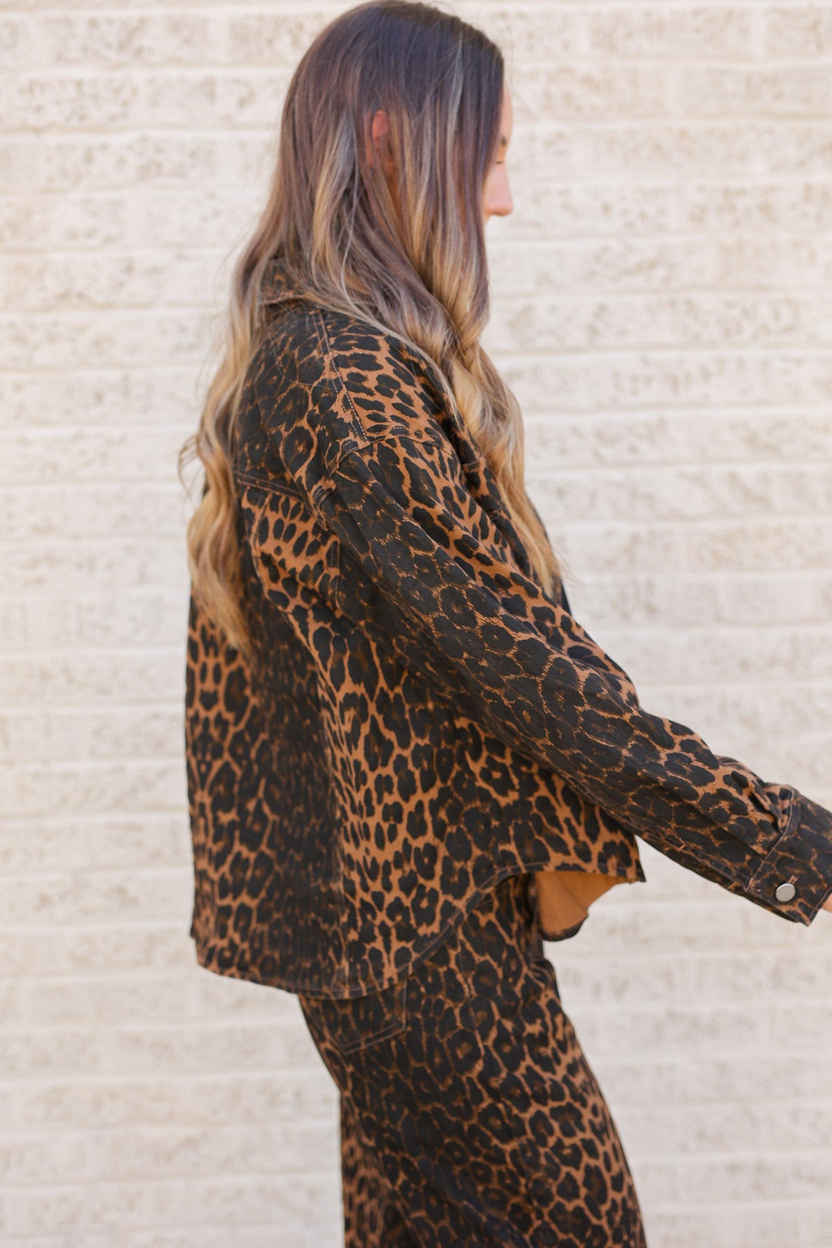 A woman shown from the side wearing a leopard-print jacket and matching pants against a light beige brick wall.
