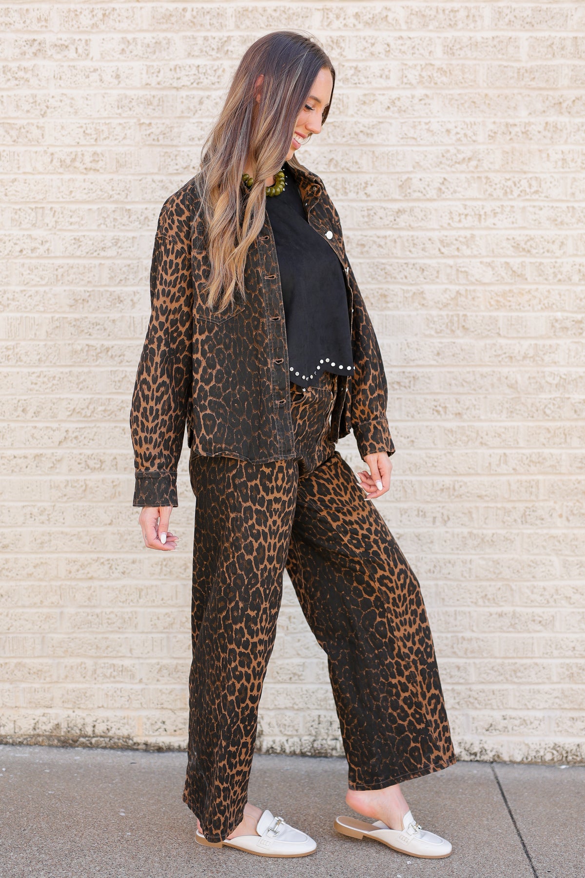 A woman wearing a matching leopard-print jacket and wide-leg pants set with a black top underneath, standing against a light beige brick wall.