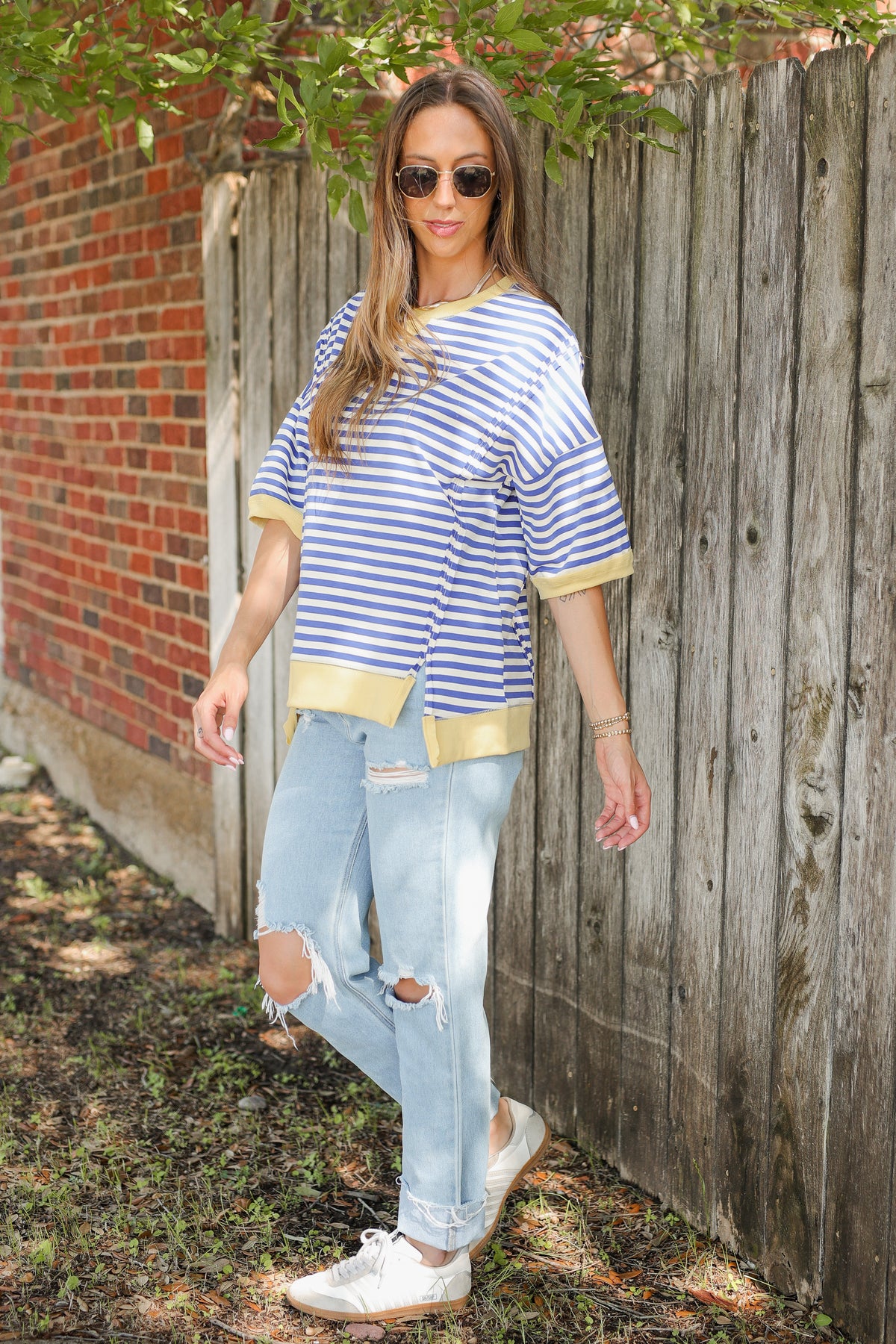 Woman wearing a blue and white striped shirt with gold accents and ripped jeans standing against a wooden fence.