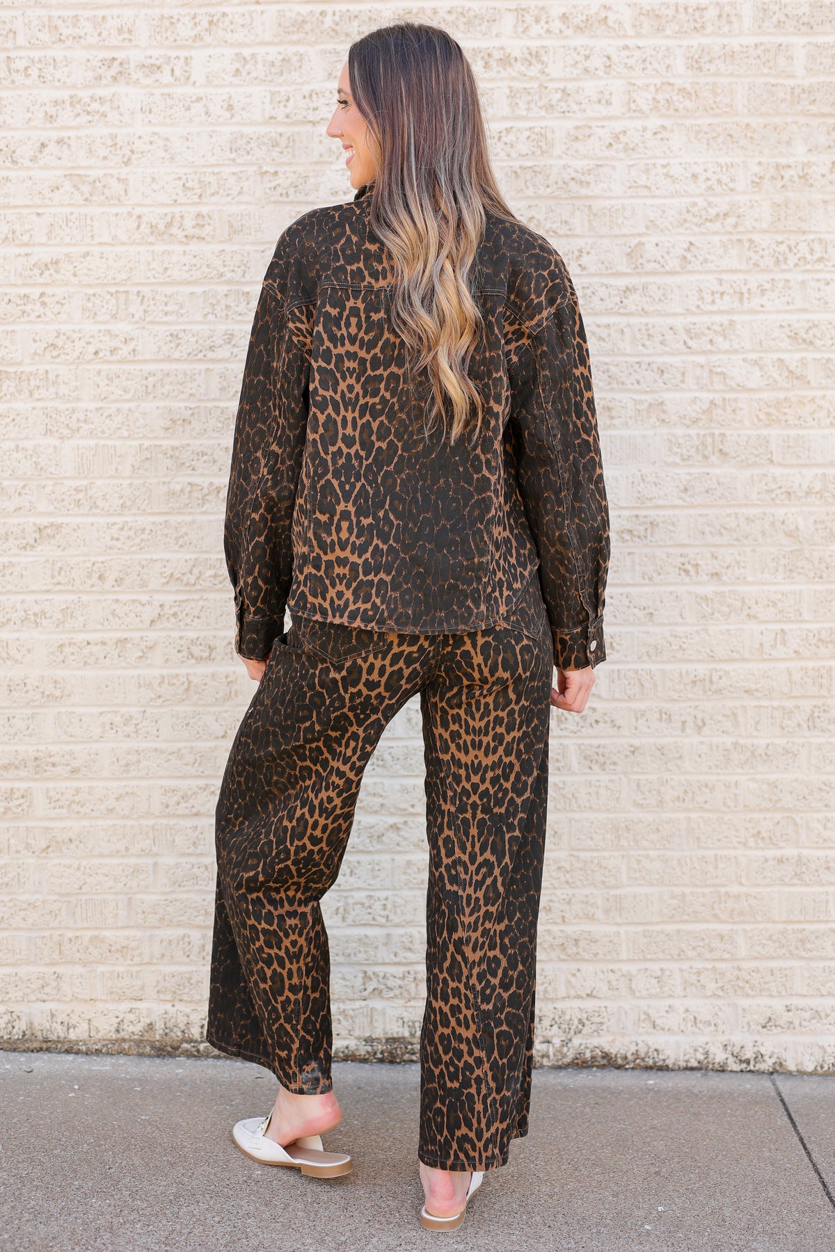 A woman stands with her back to the camera wearing a matching two-piece leopard print lounge set consisting of a long-sleeve top and wide-leg pants, paired with white slide sandals, set against a light beige brick wall.
