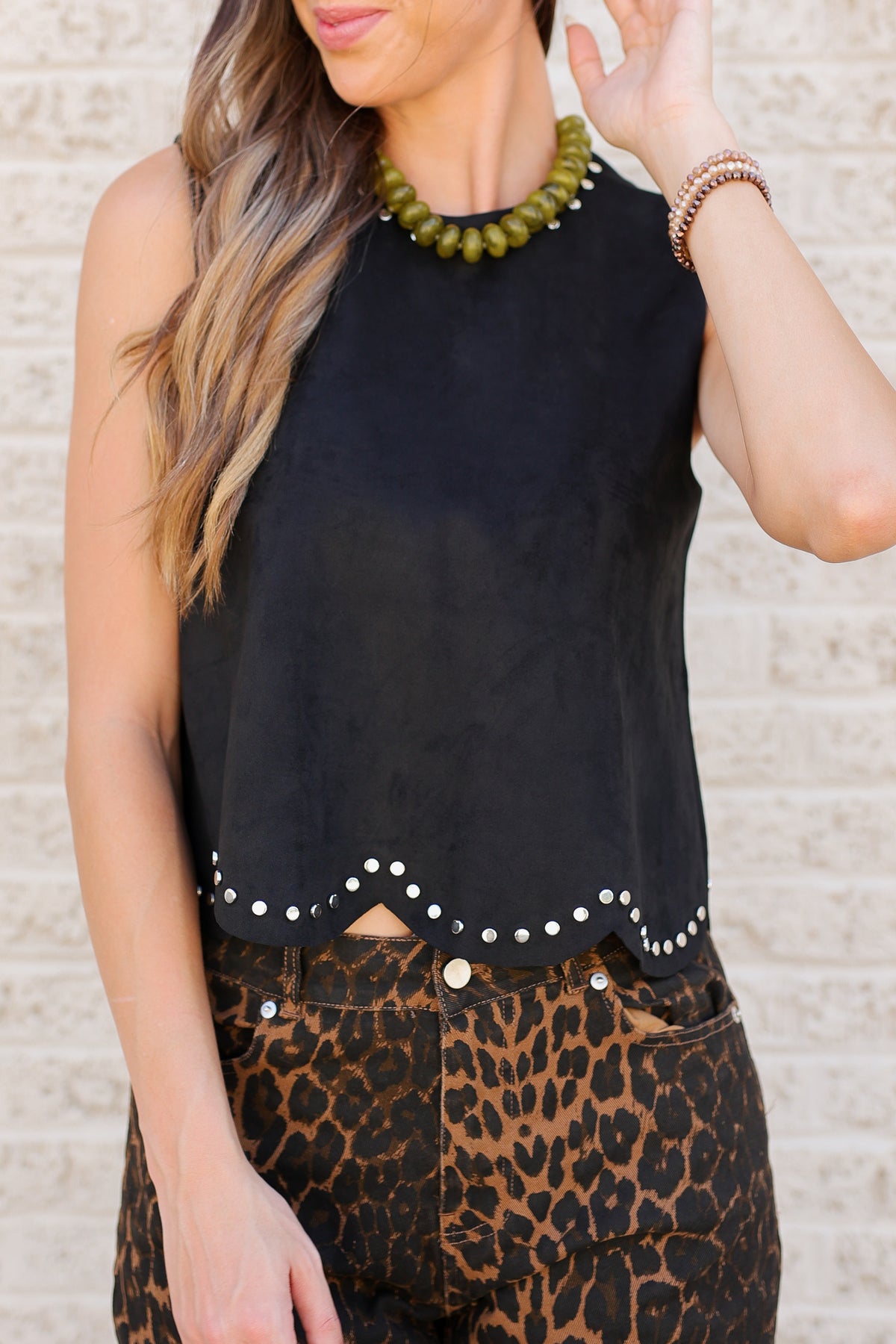 A woman wearing a sleeveless black top with a scalloped hem and white stud embellishments, paired with leopard print pants, standing against a light brick wall.