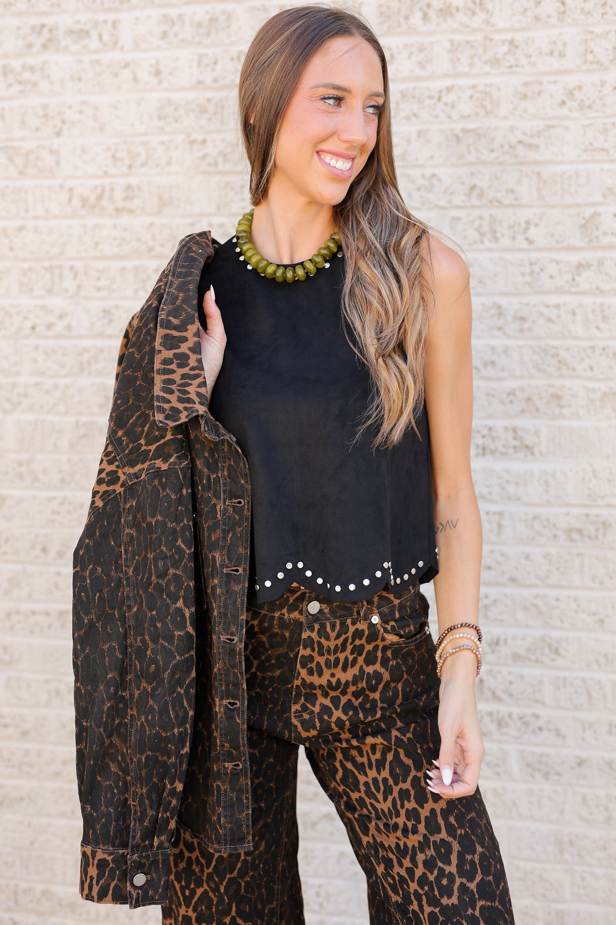 A woman wearing a black sleeveless top with silver studs, leopard-print pants, and a matching leopard-print jacket draped over her shoulder, standing against a light brick wall.