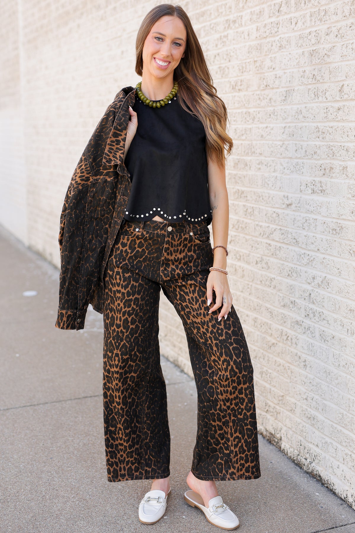 A woman stands outdoors against a light brick wall wearing a matching leopard-print blazer and wide-leg pants set with a black scalloped top and white loafers.