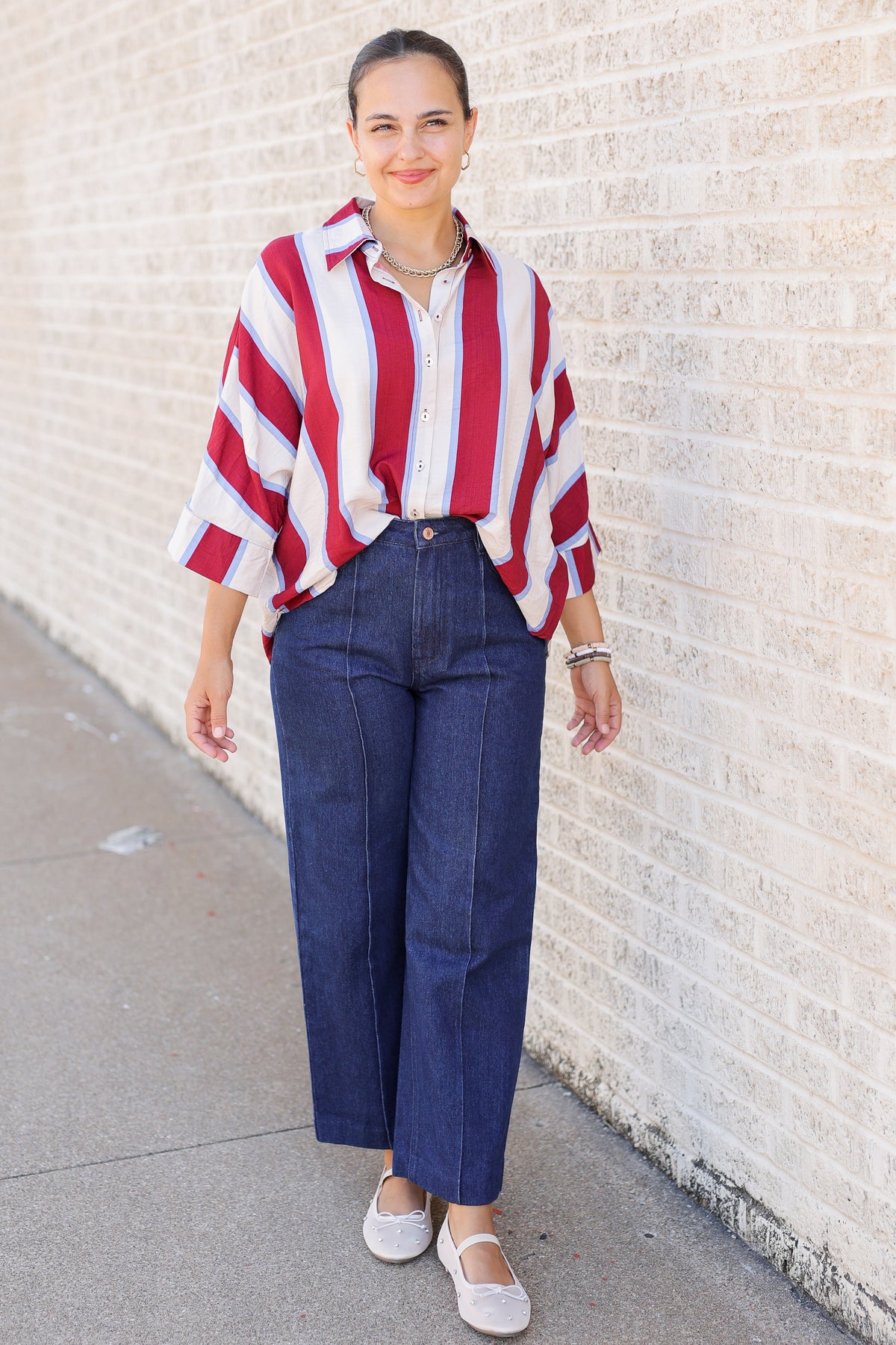 A woman stands outdoors against a light brick wall wearing a red, white, and blue vertically striped button-up shirt, dark blue wide-leg jeans, and white flats.