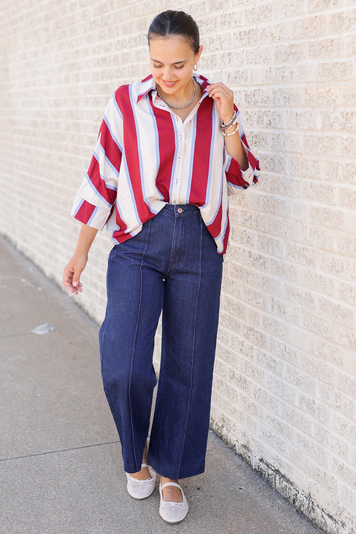 A woman stands against a light brick wall wearing a red, white, and blue vertically striped button-down shirt, dark blue wide-leg jeans, and white flats.