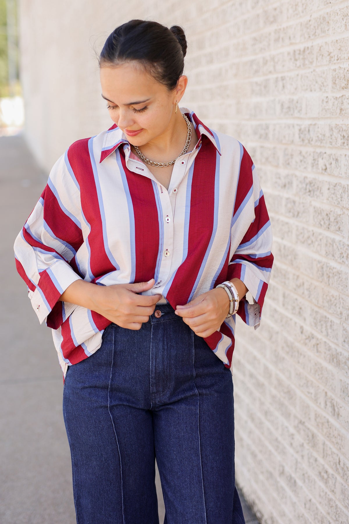 A woman wearing a red, white, and blue striped button-down shirt with dark blue jeans, standing against a light-colored brick wall.