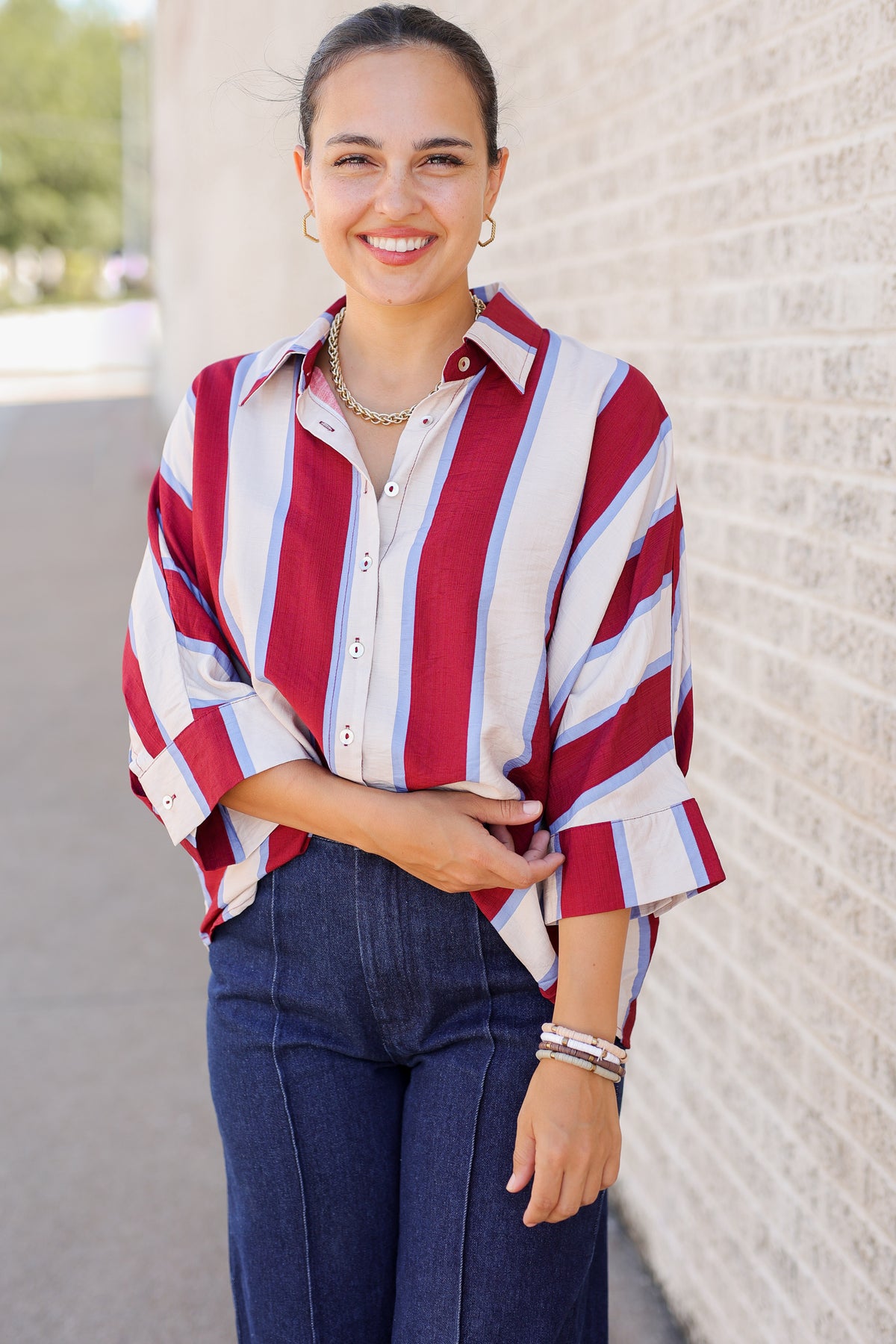 A woman smiling while wearing a red, white, and light blue vertically striped button-down shirt with dark blue jeans, standing outdoors against a light brick wall.