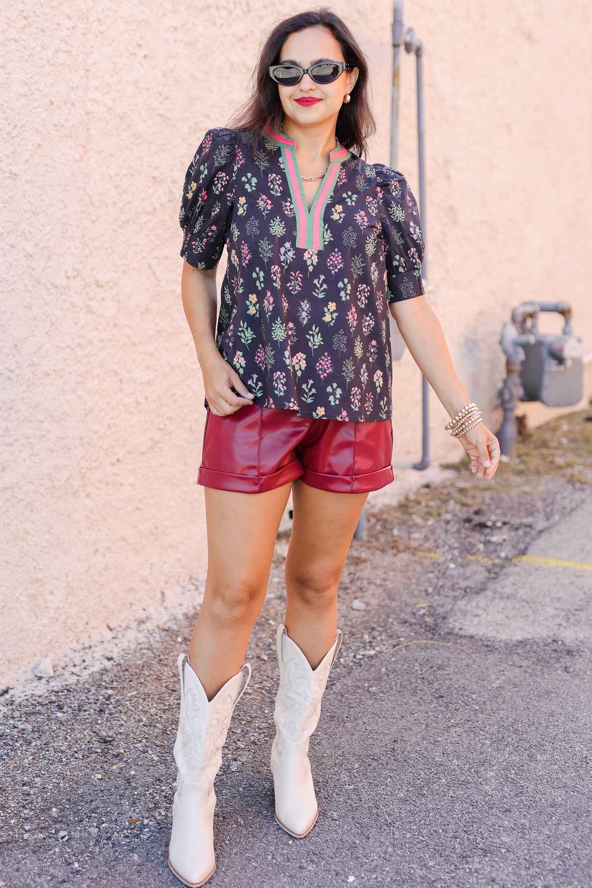 Woman wearing a floral blouse, red shorts, and white cowboy boots against a textured wall.