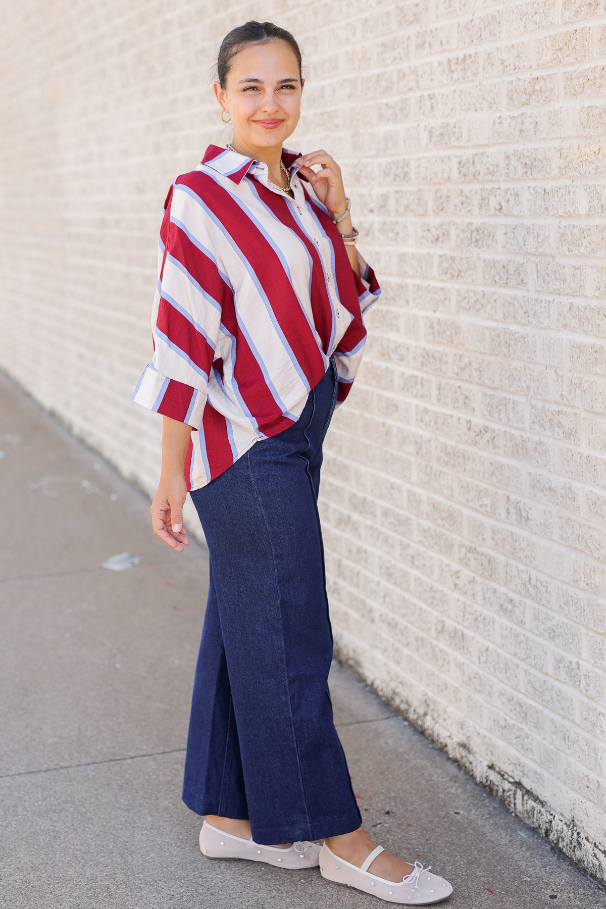 A woman stands outdoors against a light brick wall wearing a red, white, and blue striped shirt with navy wide-leg pants and white flats.