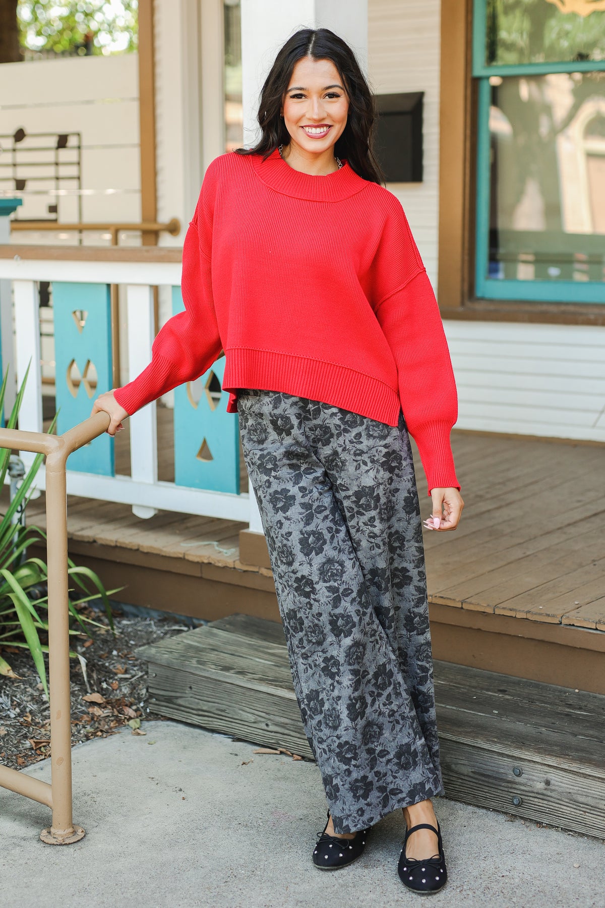 Woman wearing a red sweater and floral pants standing on a porch.