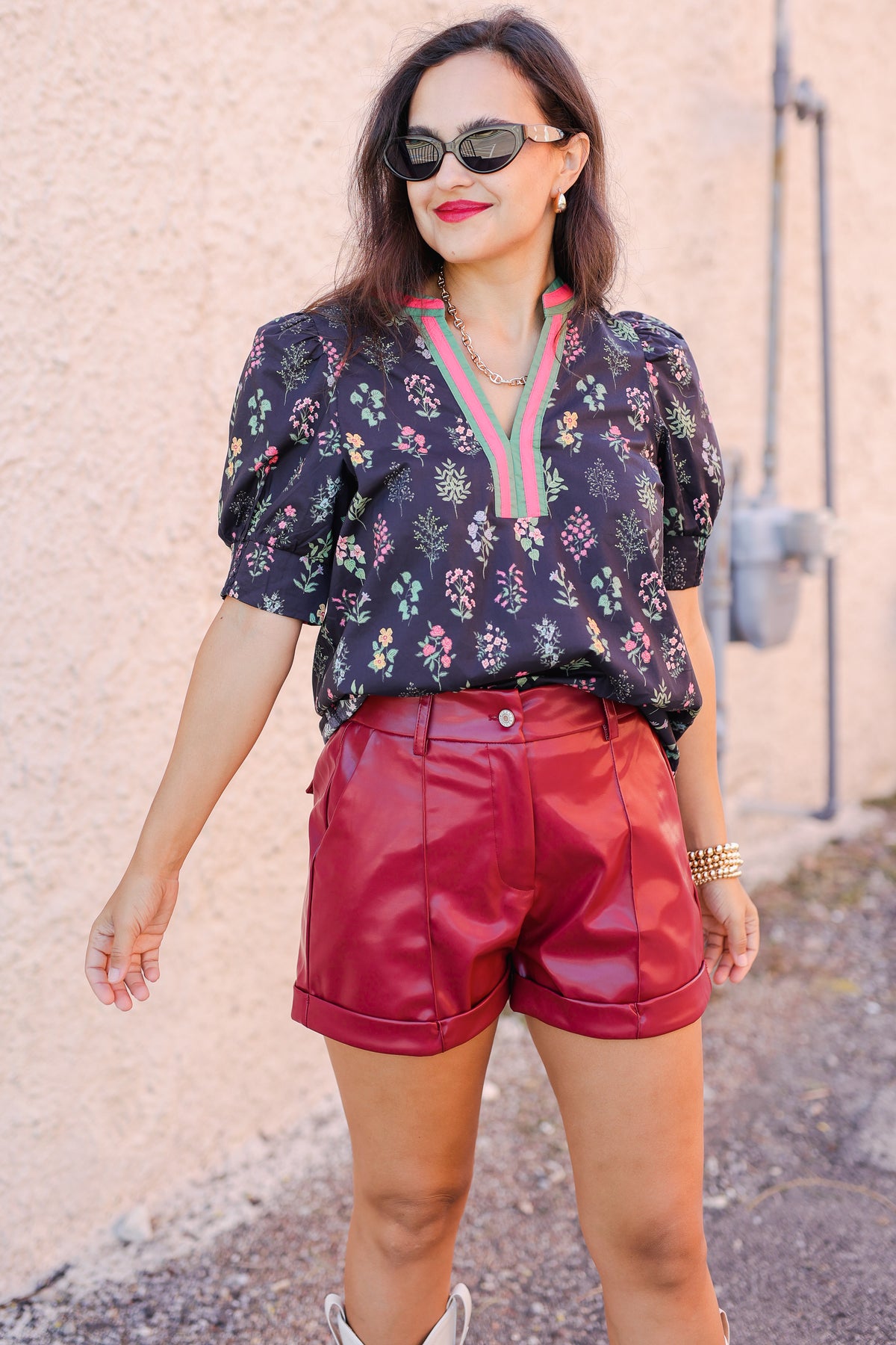 Woman wearing a floral blouse and red shorts against a textured wall.