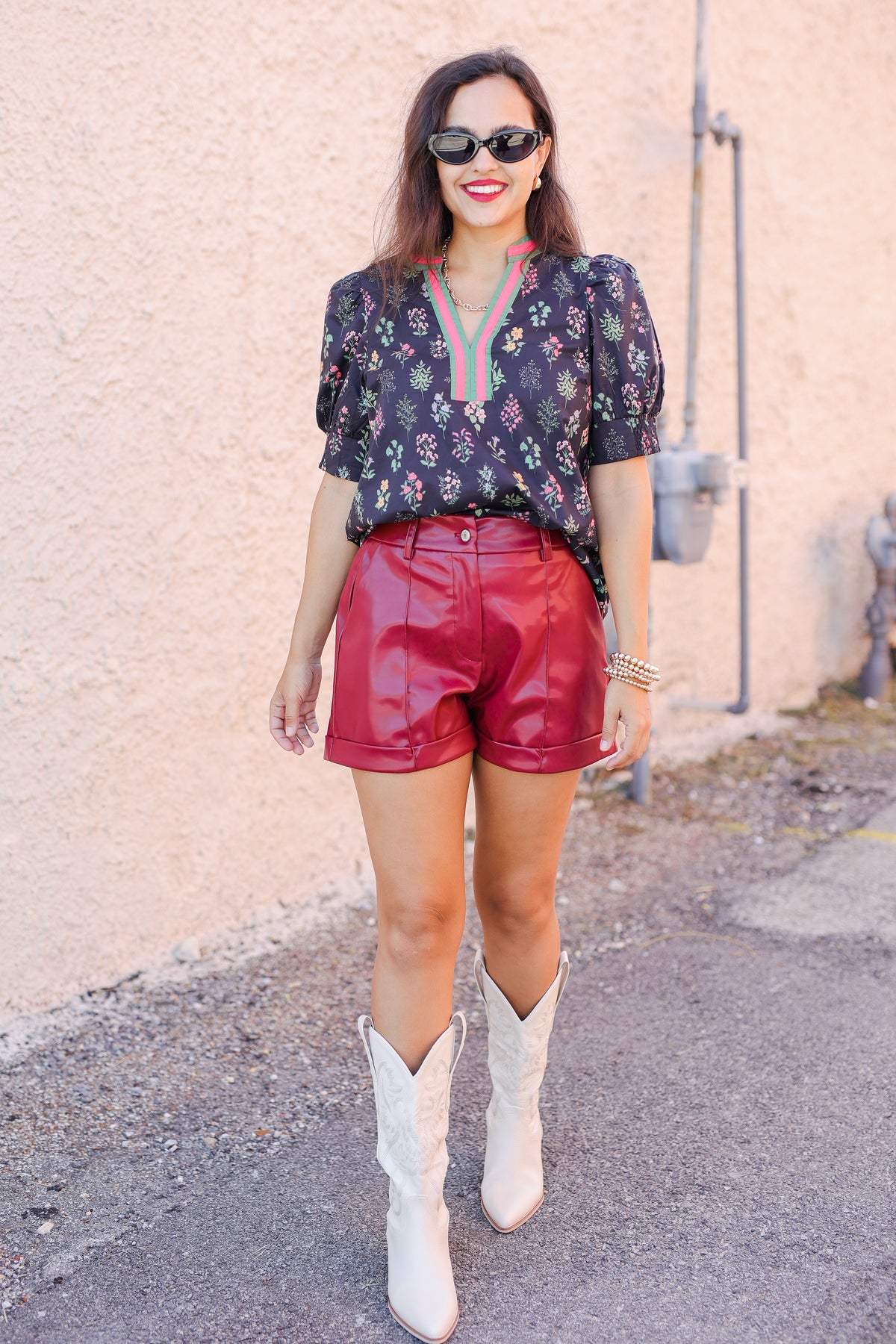 Woman wearing a floral blouse, red shorts, and white cowboy boots against a beige wall.