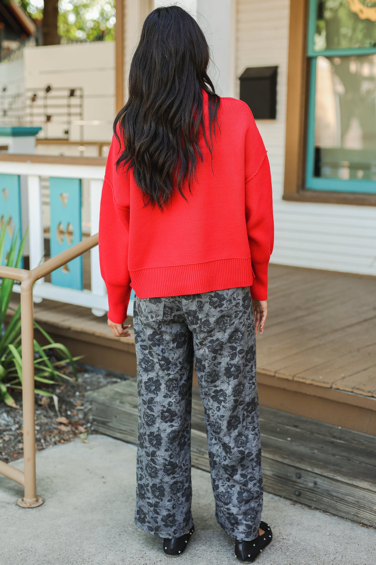 Person wearing a red sweater and patterned pants standing on a porch.