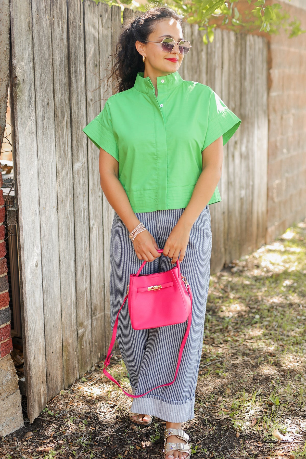 Woman in green shirt and gray pants holding a pink bag against a wooden fence.