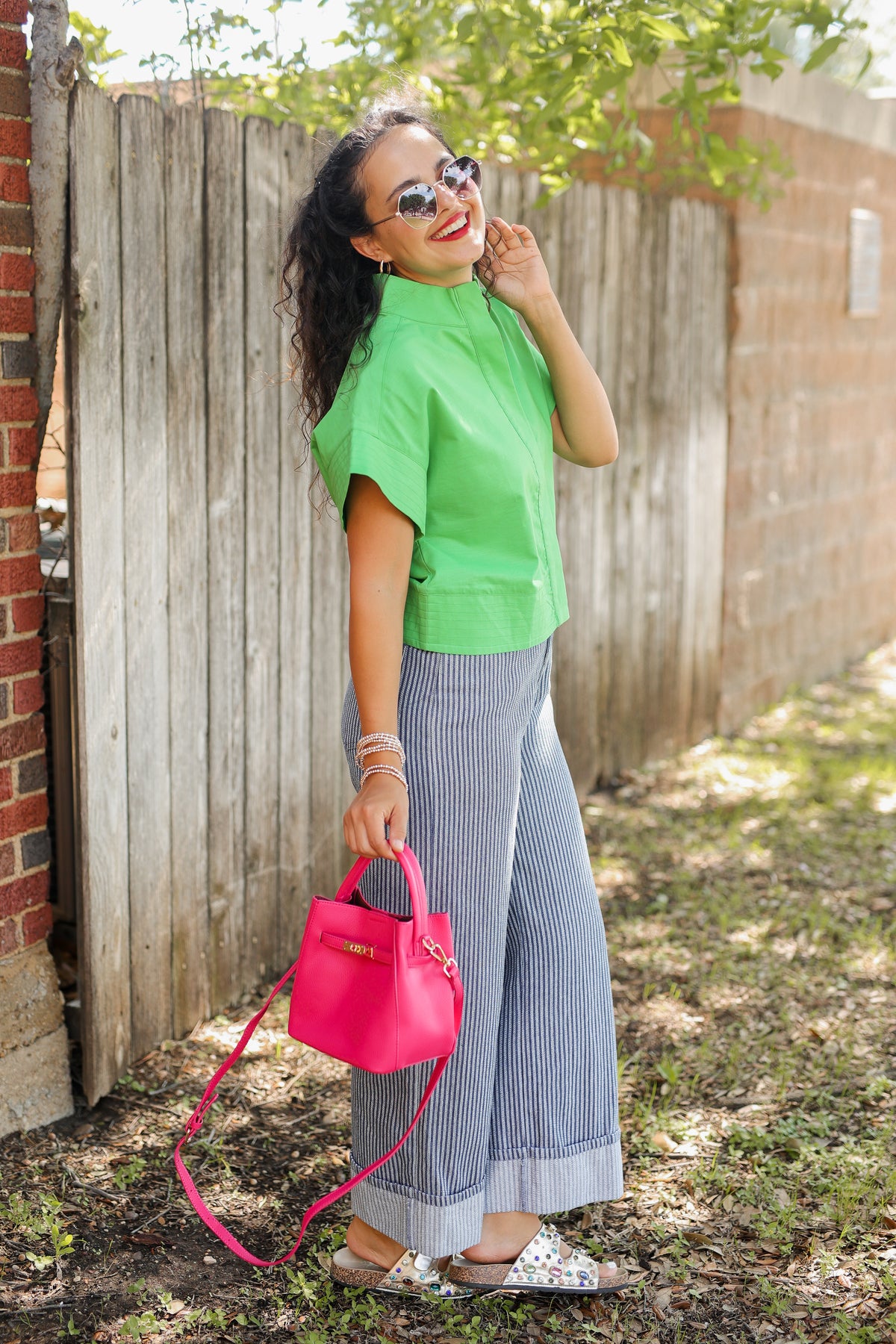 Woman in green top and blue pants holding a pink handbag outdoors.
