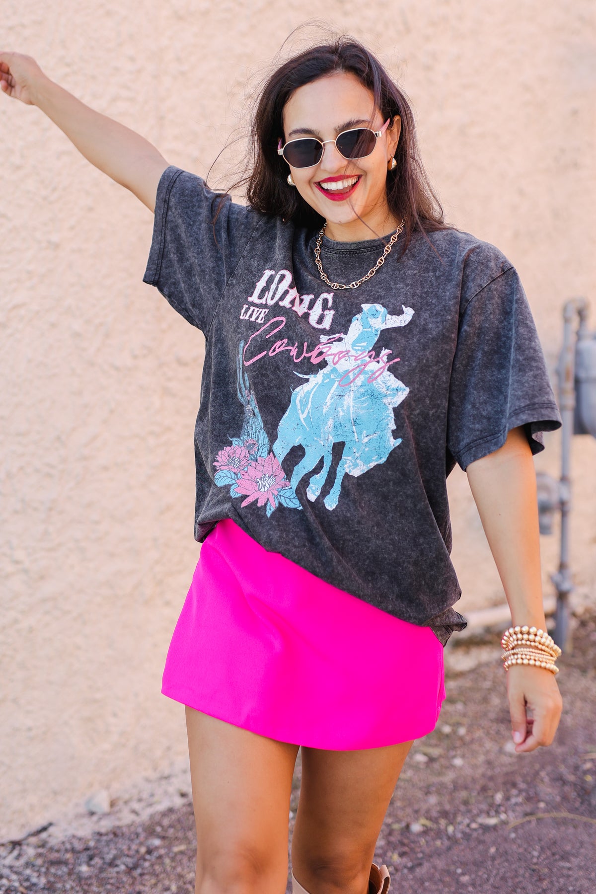 Woman wearing a graphic t-shirt and bright pink skirt with sunglasses, standing against a beige wall.