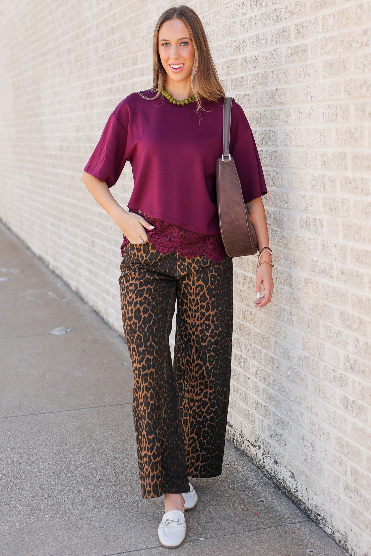 A woman stands against a light brick wall wearing a burgundy short-sleeve top, leopard-print pants, white slip-on shoes, and carrying a brown shoulder bag.