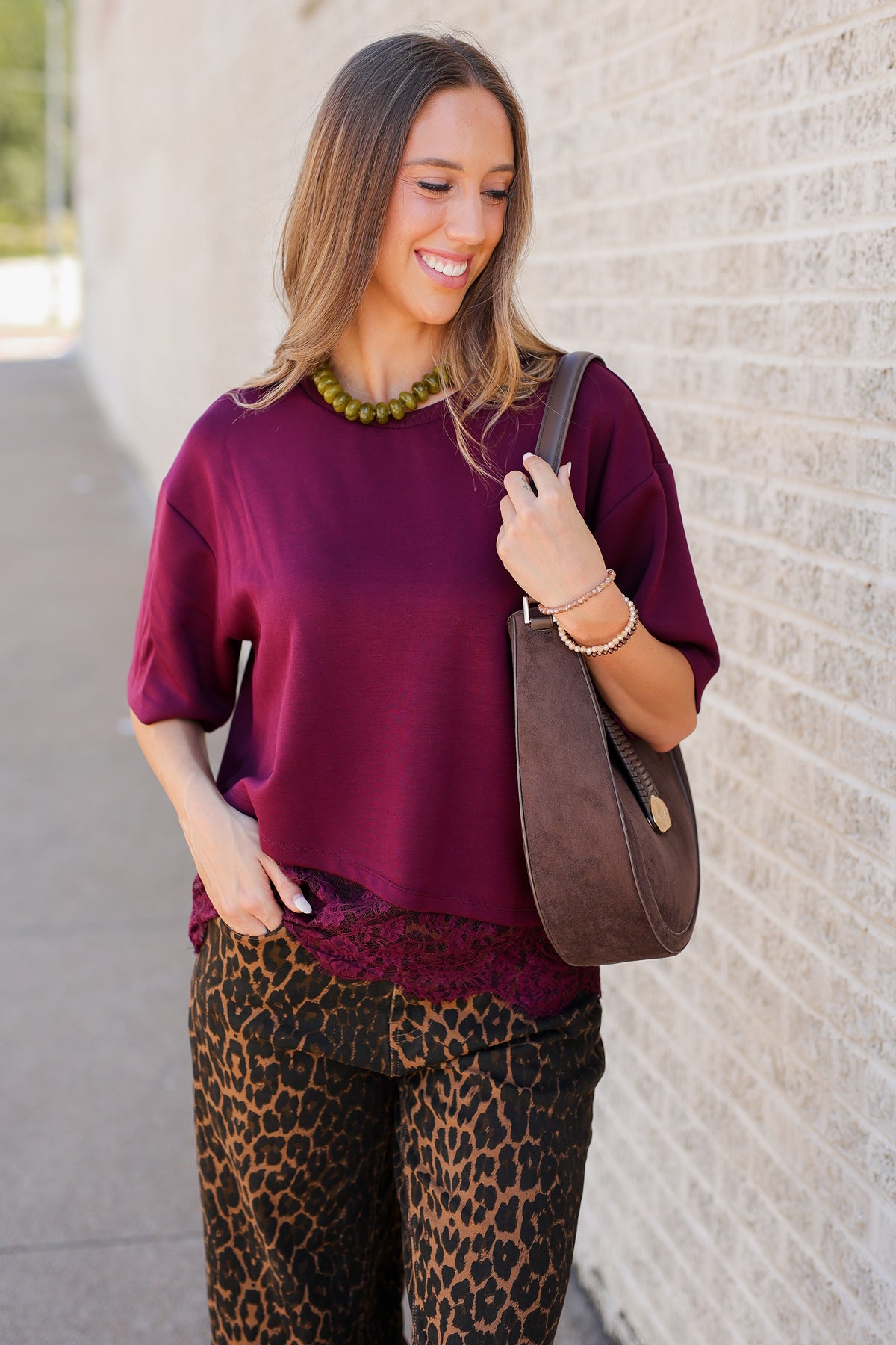 A woman wearing a loose-fitting deep burgundy top and leopard-print pants, carrying a brown shoulder bag, standing outdoors against a light brick wall.