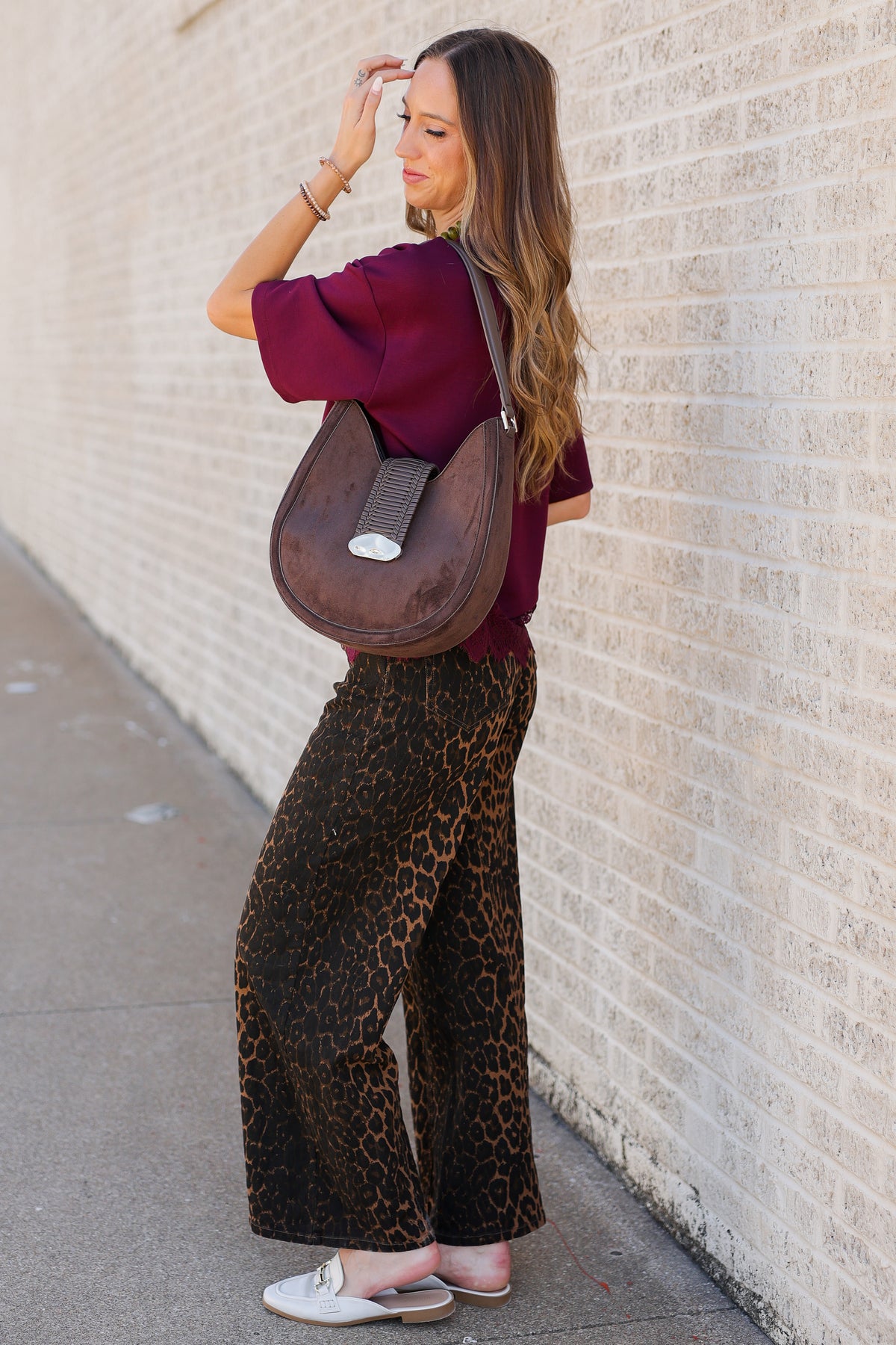 A woman stands on a sidewalk against a light brick wall, wearing a burgundy short-sleeve top, brown leopard-print wide-leg pants, and white slip-on shoes, while carrying a brown crescent-shaped shoulder bag.