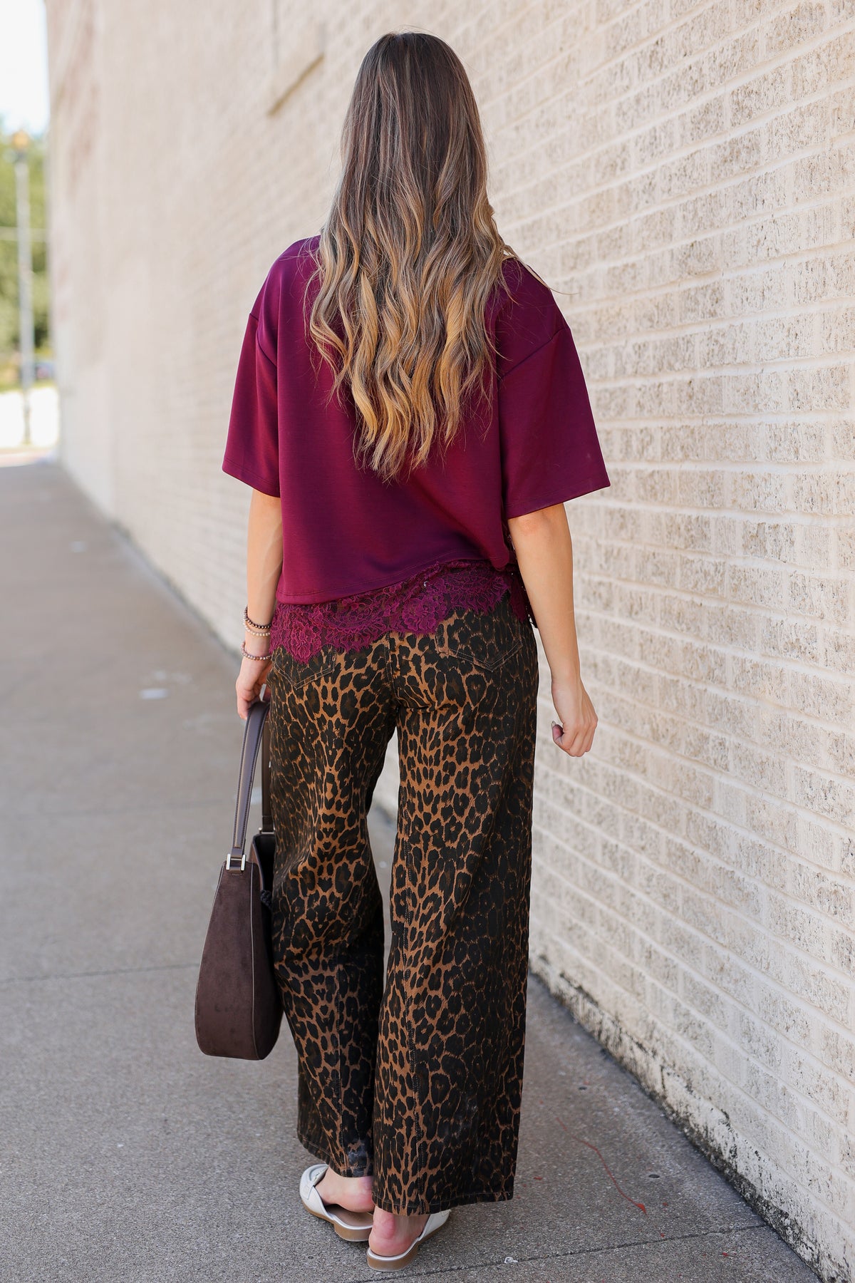 A woman viewed from behind wearing a loose burgundy t-shirt with a lace hem and wide-leg leopard-print pants, walking along a sidewalk next to a light brick wall.