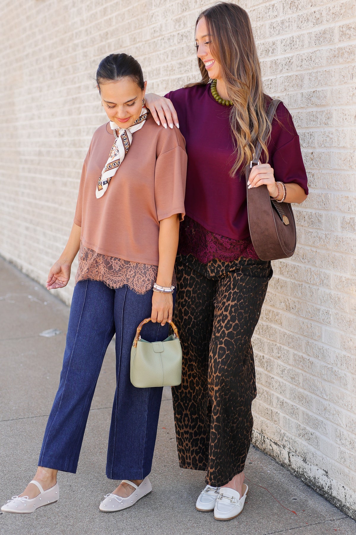 Two women posing outdoors against a light brick wall, showcasing different fashion outfits. One wears a dusty rose top with lace trim, navy wide-leg jeans, and white flats, holding a small sage green handbag.