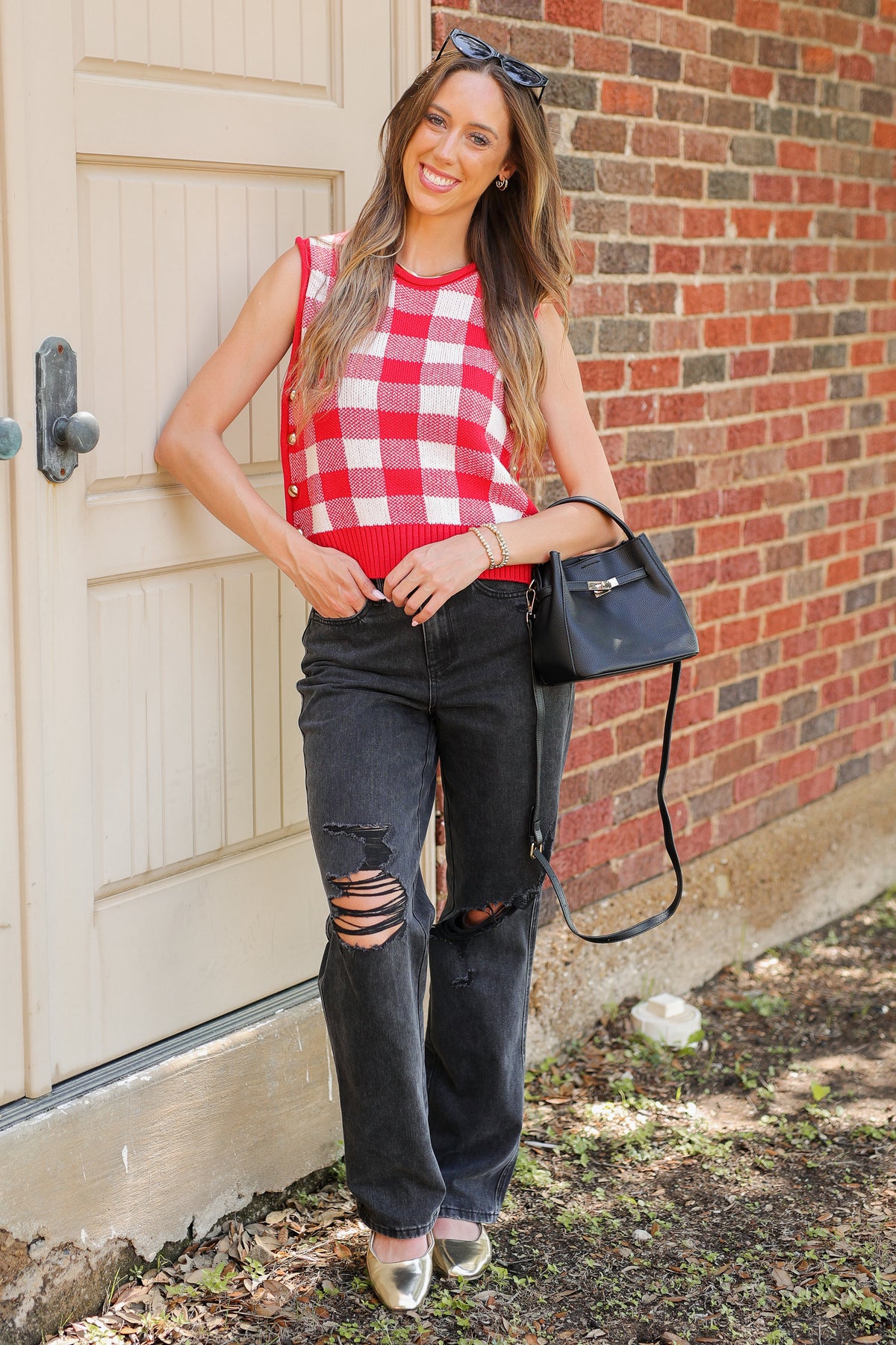 A woman stands outdoors against a brick wall and cream-colored door, wearing a red and white gingham sleeveless top, black ripped jeans, and carrying a black shoulder bag.