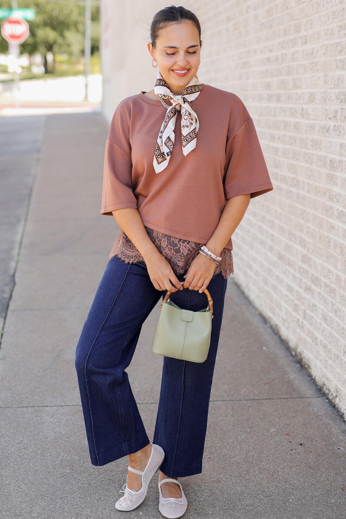 A woman stands on a sidewalk wearing a dusty rose short-sleeve top with a patterned neck scarf, navy wide-leg pants, white flats, and holding a sage green handbag.