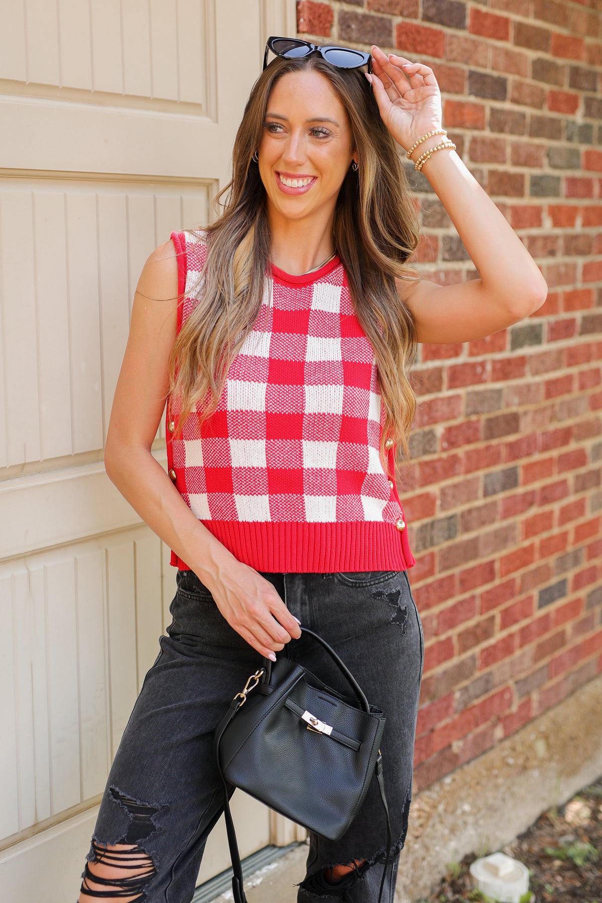 A woman wearing a red and white gingham knit sleeveless top, black ripped jeans, and carrying a black handbag, posing in front of a brick wall and garage door.