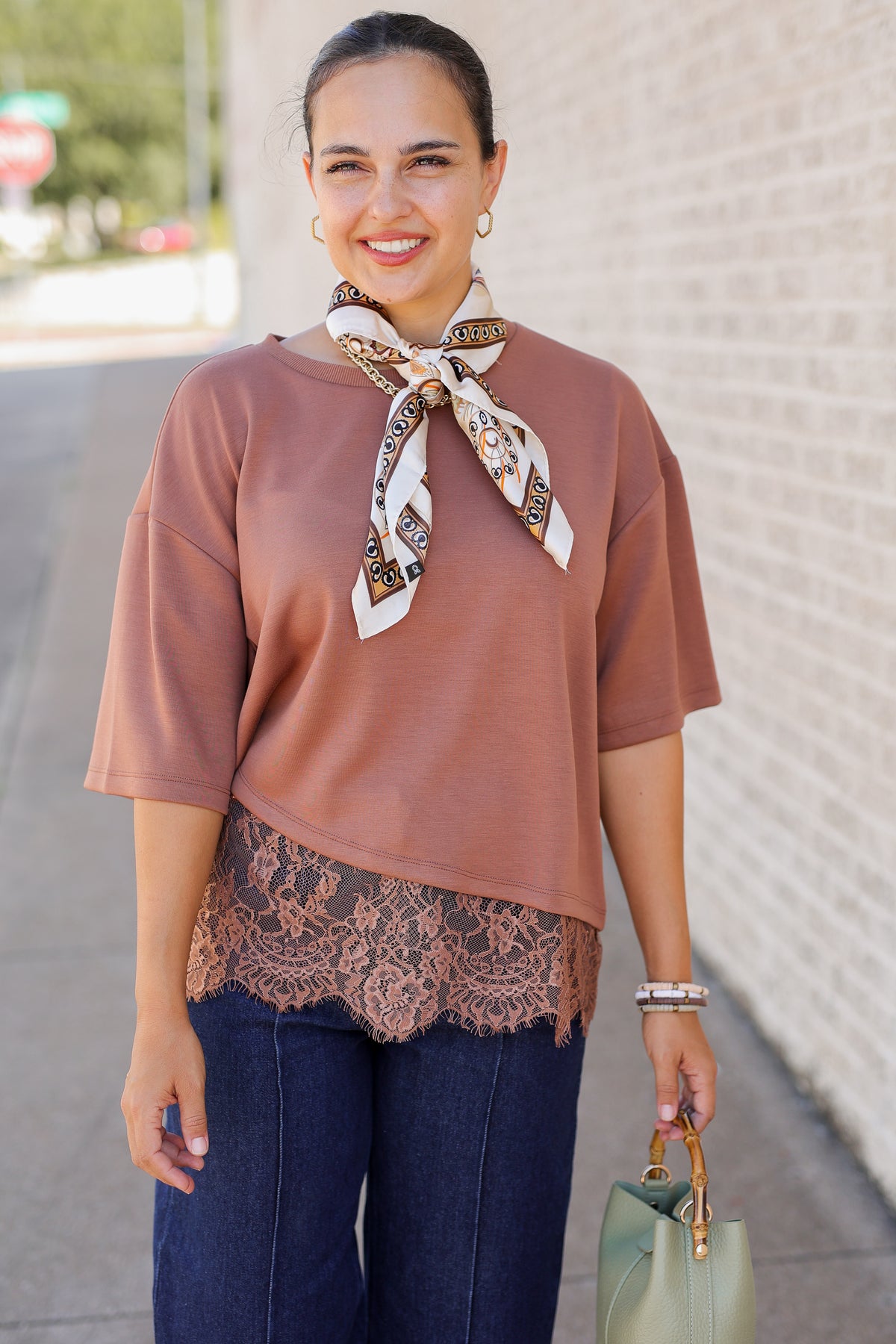 A woman wearing a dusty rose short-sleeve top with a lace hem, styled with a patterned neck scarf, dark blue jeans, and holding a sage green handbag, standing outdoors against a light brick wall.
