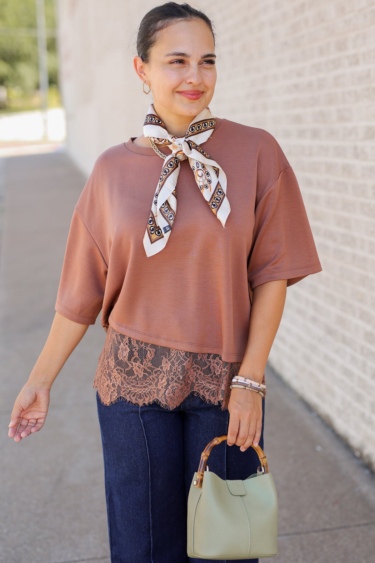A woman wearing a loose, short-sleeve dusty rose-brown top with a patterned neck scarf, dark lace hem detail, dark blue jeans, and holding a sage green handbag, standing outdoors against a light brick wall.