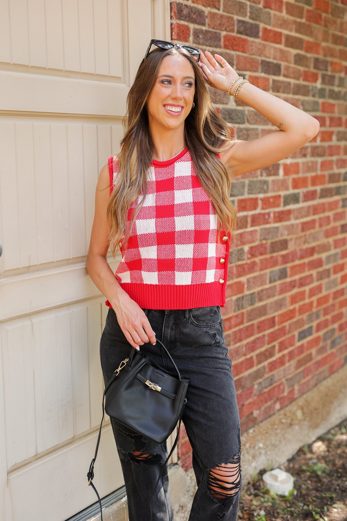 Woman wearing a red and white checkered sleeveless top with black ripped jeans, standing against a brick wall.