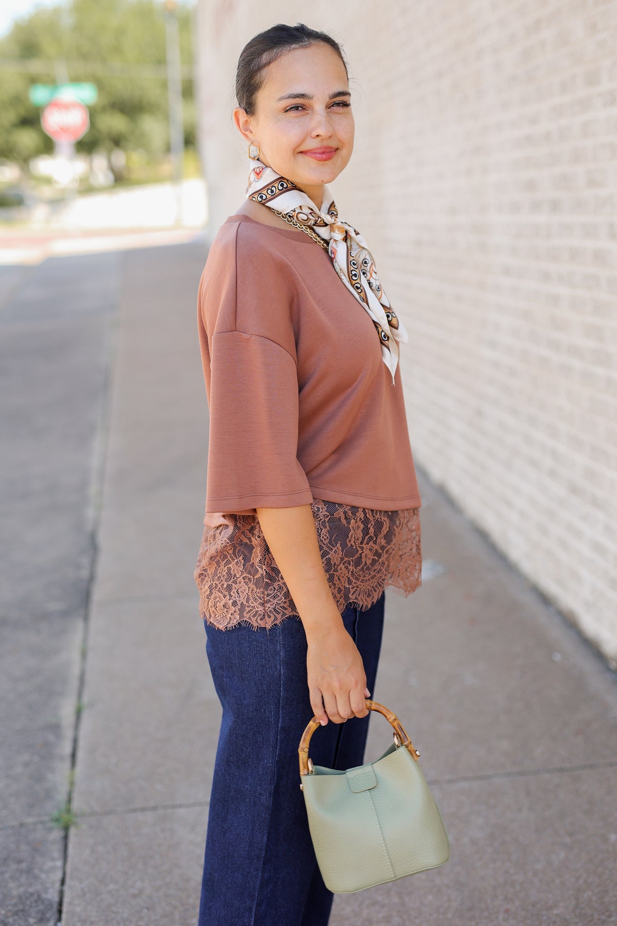 A woman stands on a sidewalk wearing a dusty rose short-sleeve top with a lace hem, a patterned neck scarf, dark blue jeans, and holding a sage green handbag with a bamboo handle.