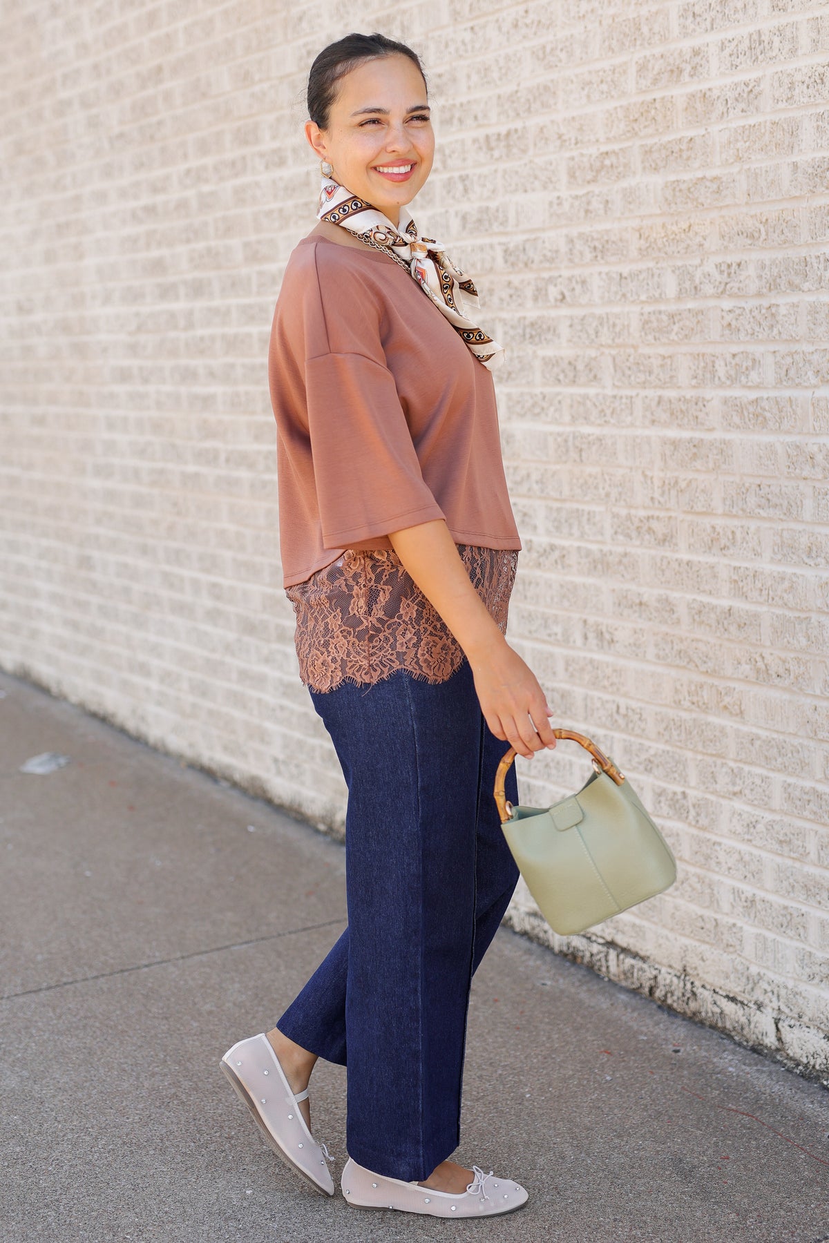 A woman posing outdoors against a light brick wall, wearing a dusty rose short-sleeve top with a lace hem, dark blue wide-leg jeans, white slip-on shoes, and holding a sage green handbag.