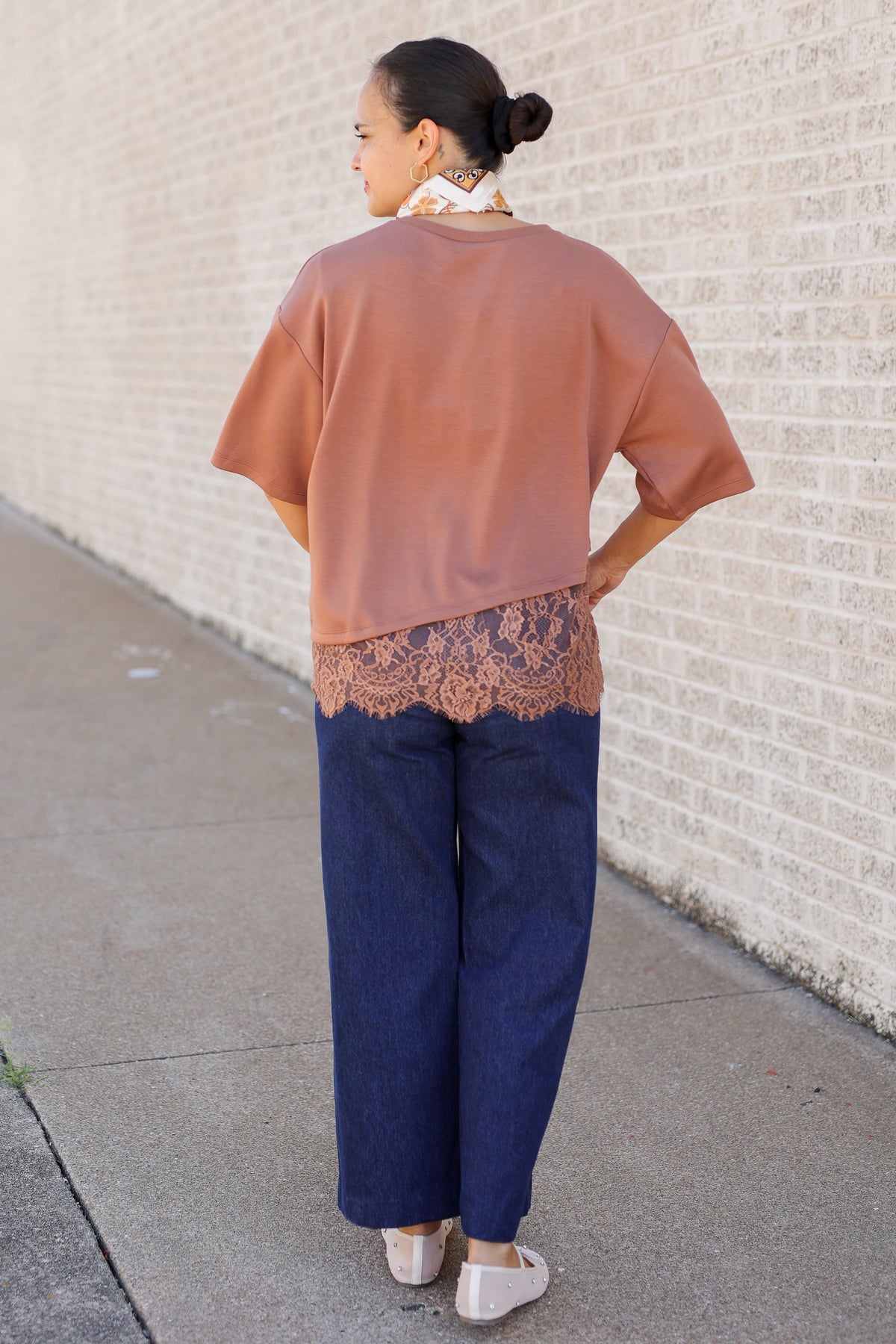 A woman stands on a sidewalk against a light brick wall, viewed from behind. She is wearing a loose dusty rose short-sleeve top with a lace band detail at the hem, dark blue wide-leg pants, and white shoes.