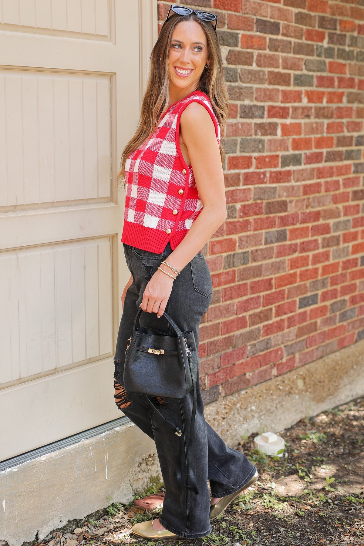 A woman stands outdoors against a brick wall and garage door wearing a red and white gingham sleeveless top, dark gray jeans, and carrying a black handbag.
