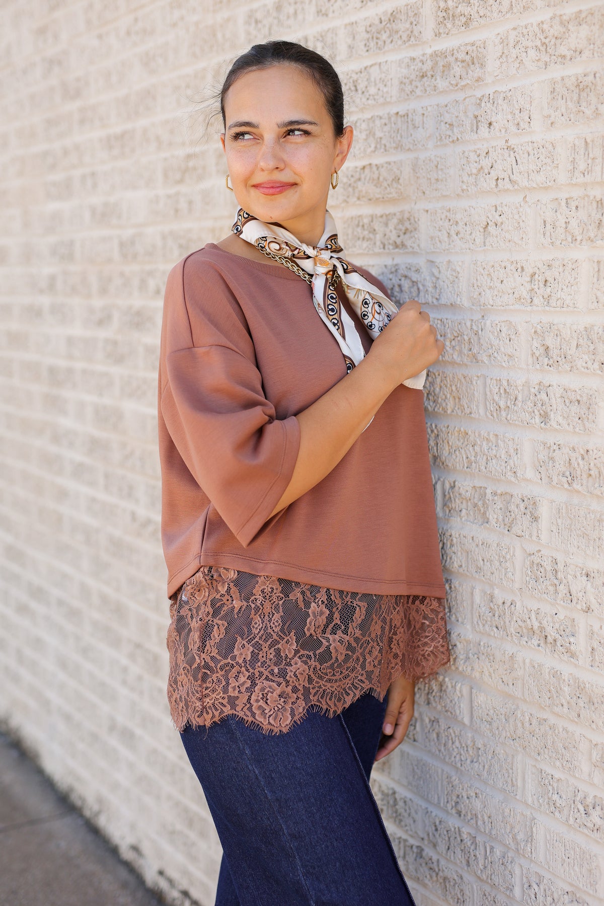 A woman wearing a dusty rose-brown blouse with a lace hem and a patterned neck scarf, leaning against a light-colored brick wall.