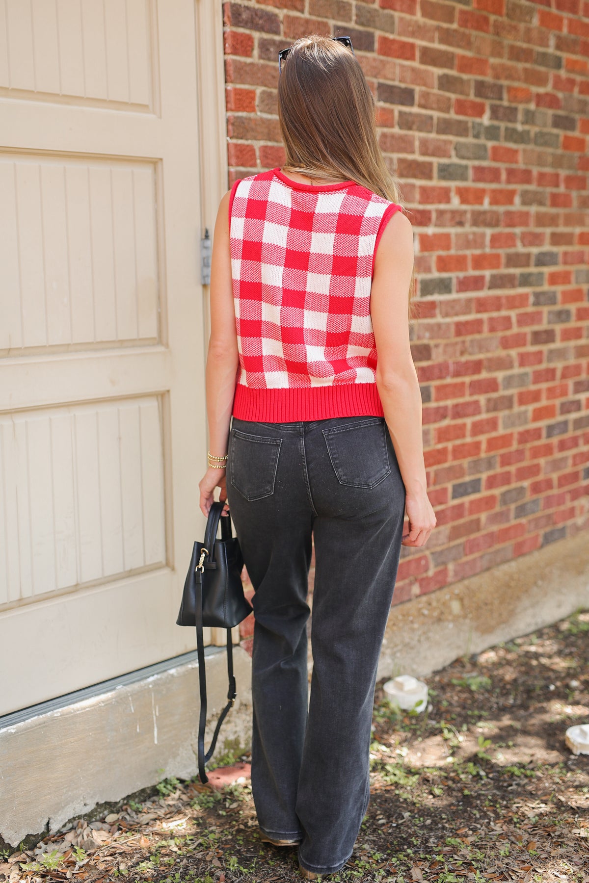A woman viewed from behind wearing a red and white gingham sleeveless knit top, charcoal gray wide-leg jeans, and holding a black handbag, standing outdoors next to a brick wall and a cream-colored door.